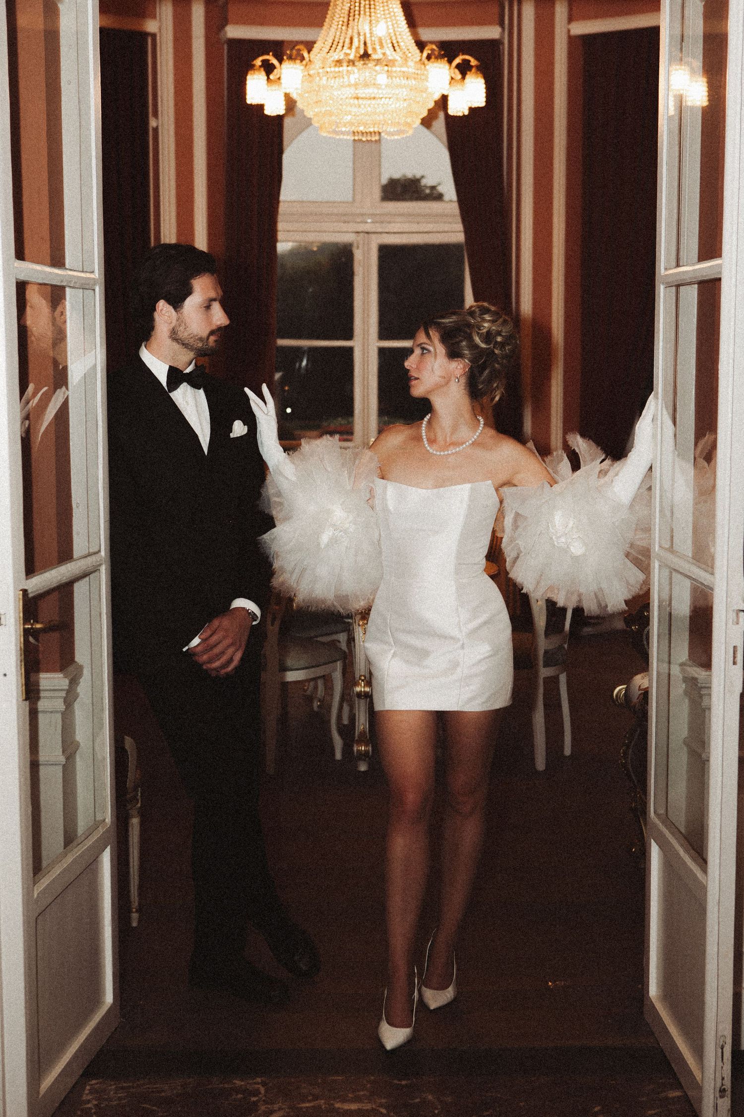 Elegant bride in white mini dress with feather sleeves and groom in tuxedo standing in doorway under chandelier.