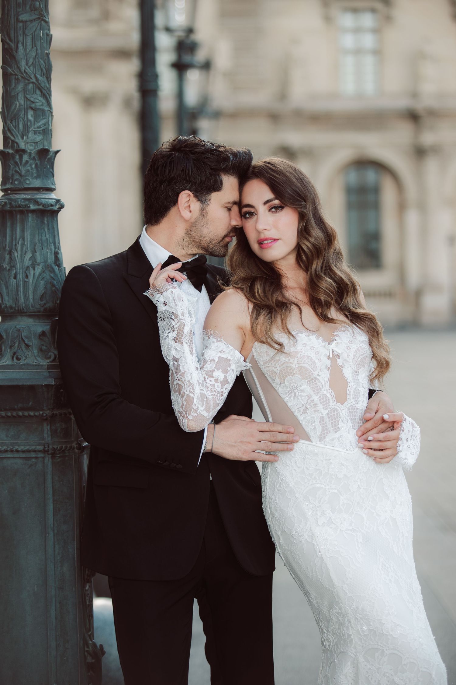 Elegant couple embraces near ornate lamppost, bride in lace off-shoulder gown and groom in black tuxedo in Paris.