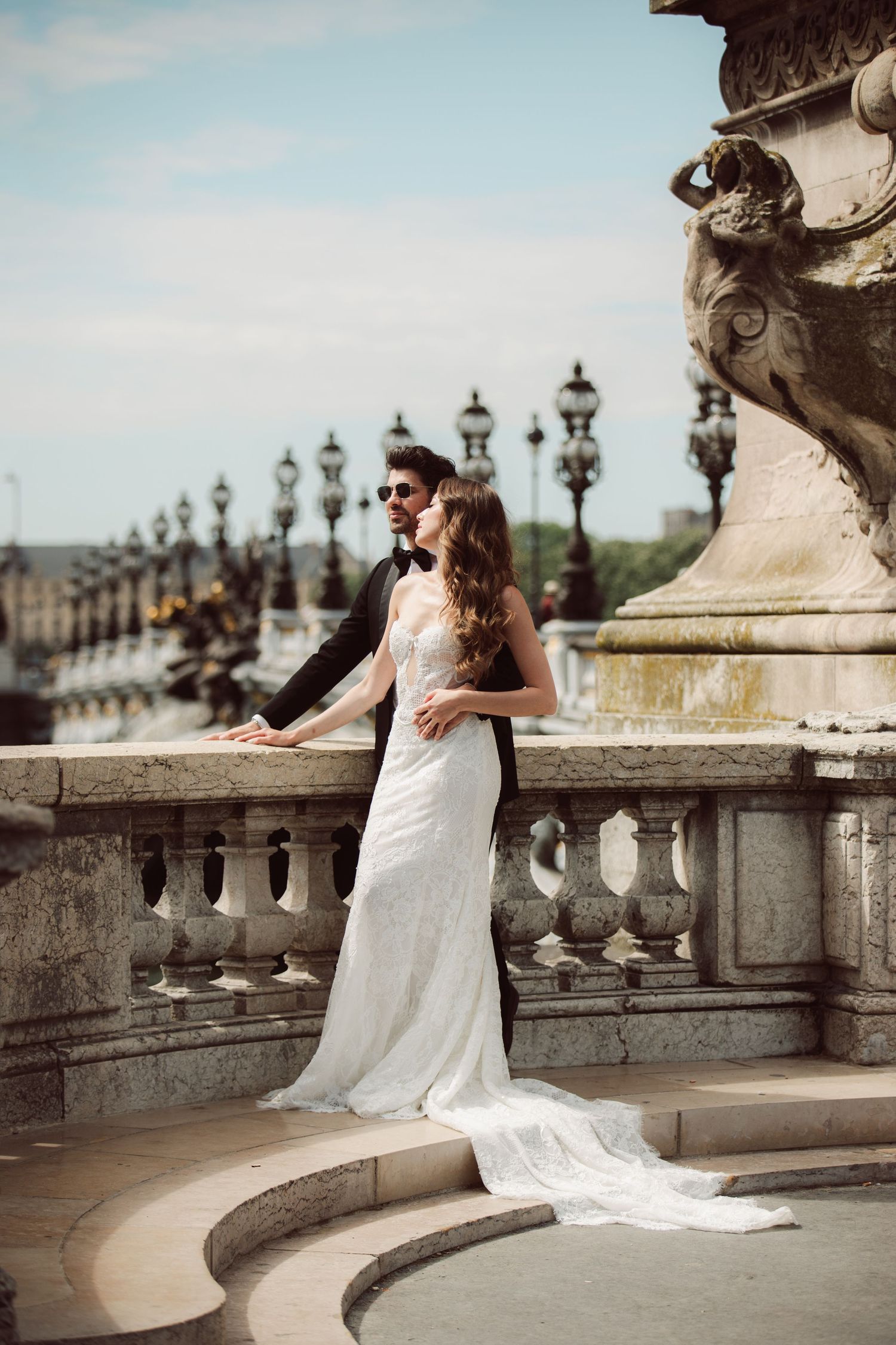 Elegant bride in a white gown and groom embrace on a ornate Parisian bridge balustrade.
