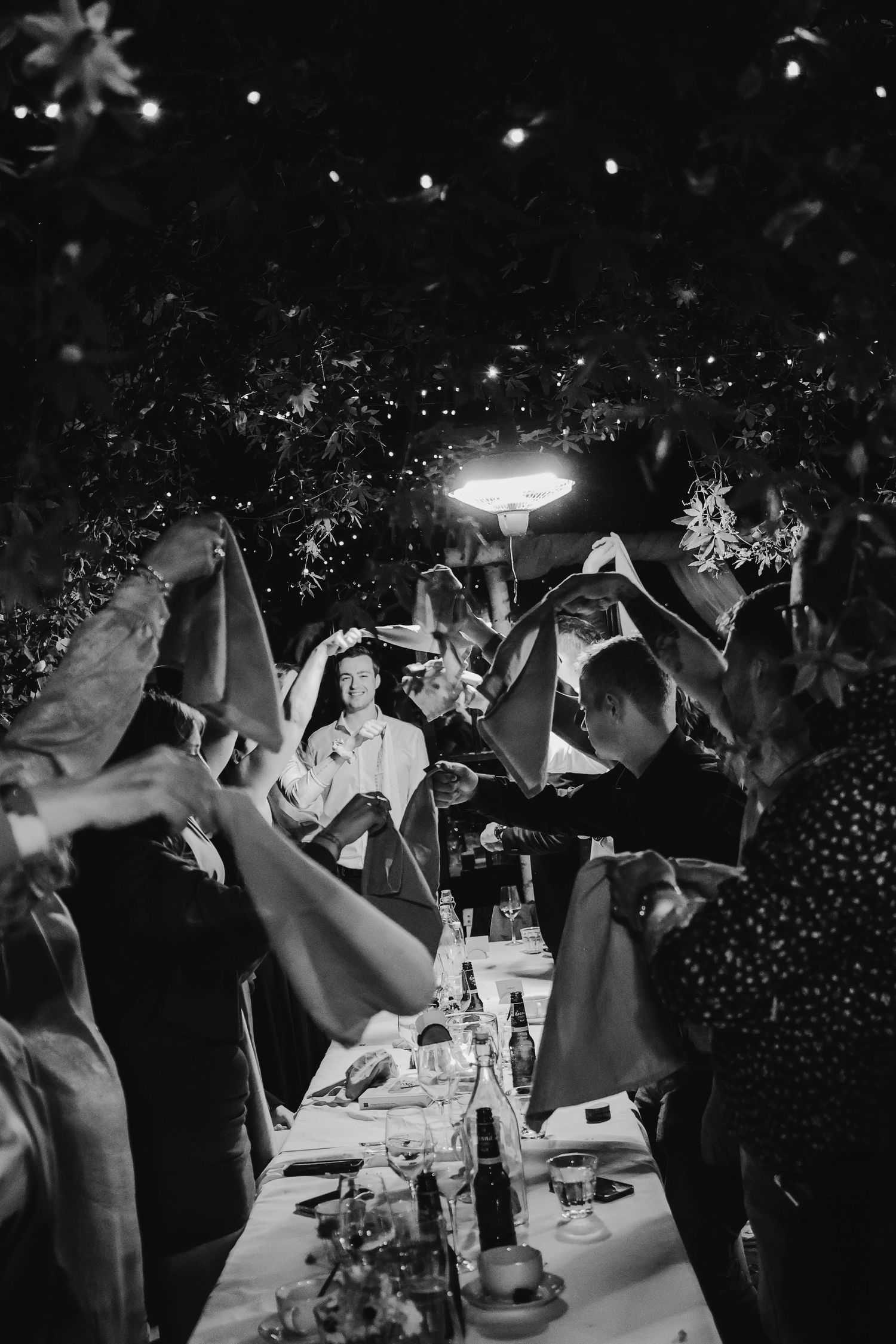 Black and white wedding reception photo of bride and groom celebrated by guests waving napkins at outdoor dinner table.