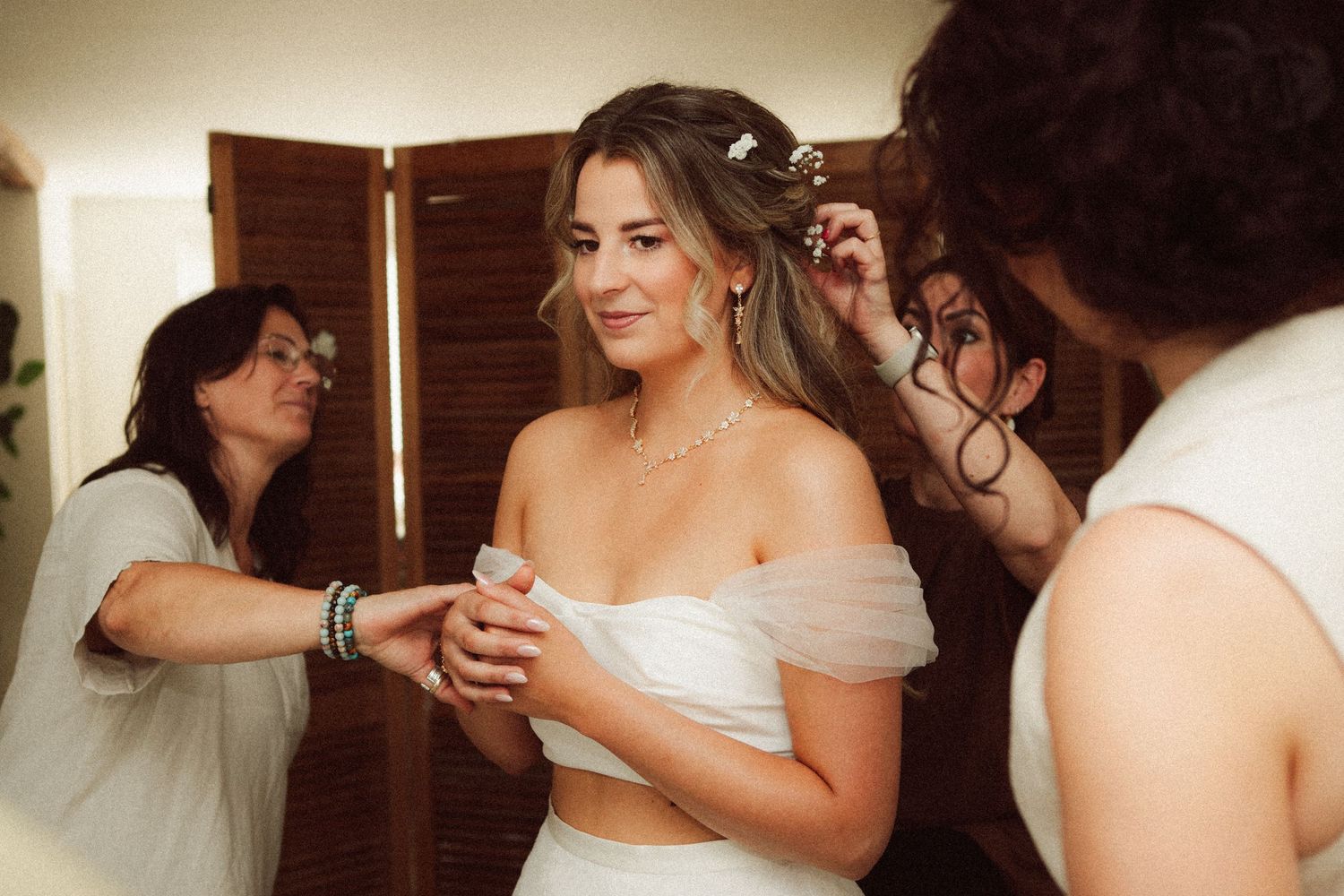 Bride smiling as two women help with her hair and dress in a bright room before her wedding.