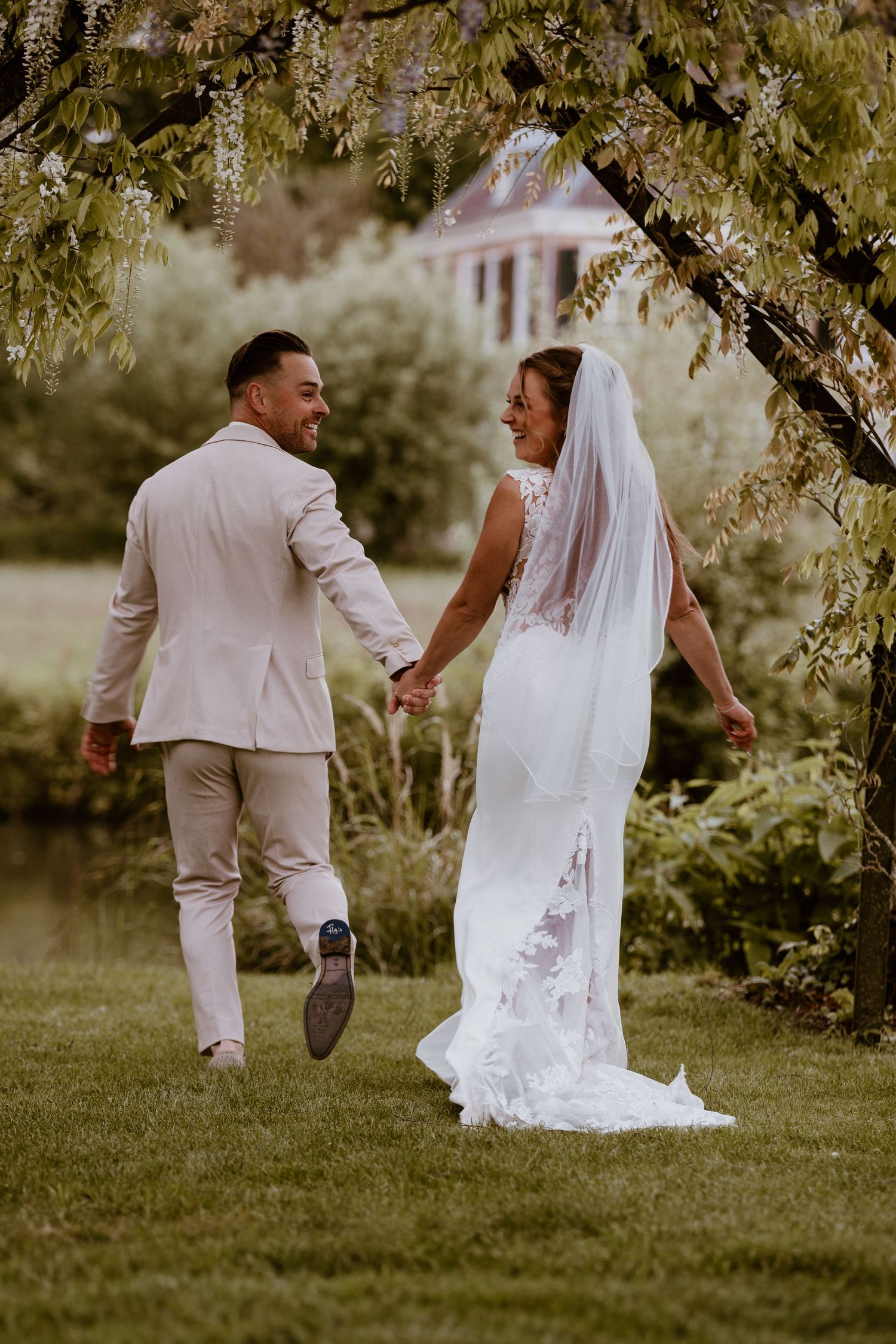 Bride in lace gown and groom in beige suit hold hands walking through a garden.