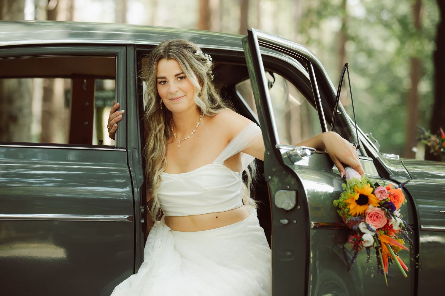 Smiling bride in two-piece wedding dress poses beside vintage dark green car in forest.