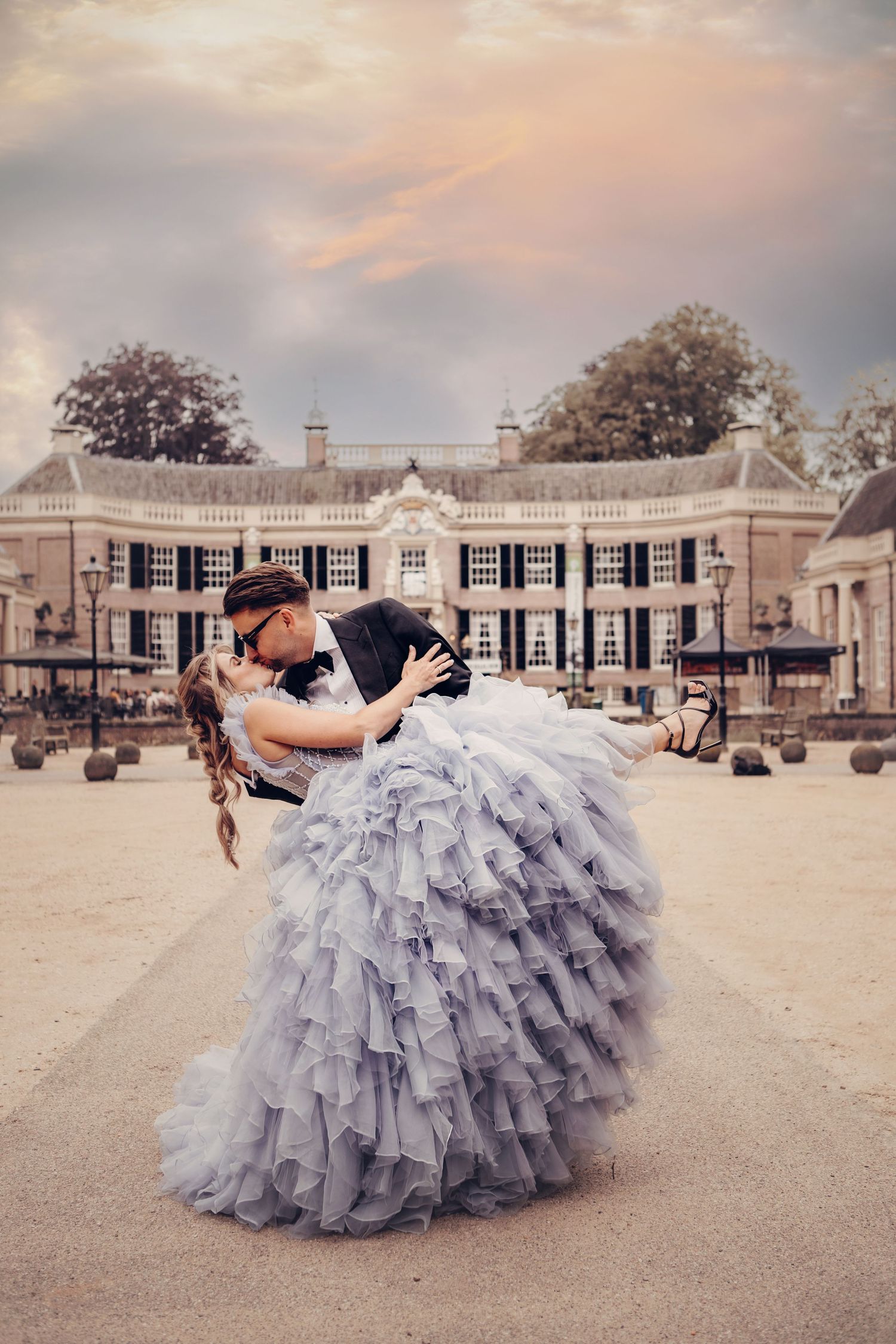 Groom dips bride in stunning grey ruffled gown outside elegant European mansion at sunset.