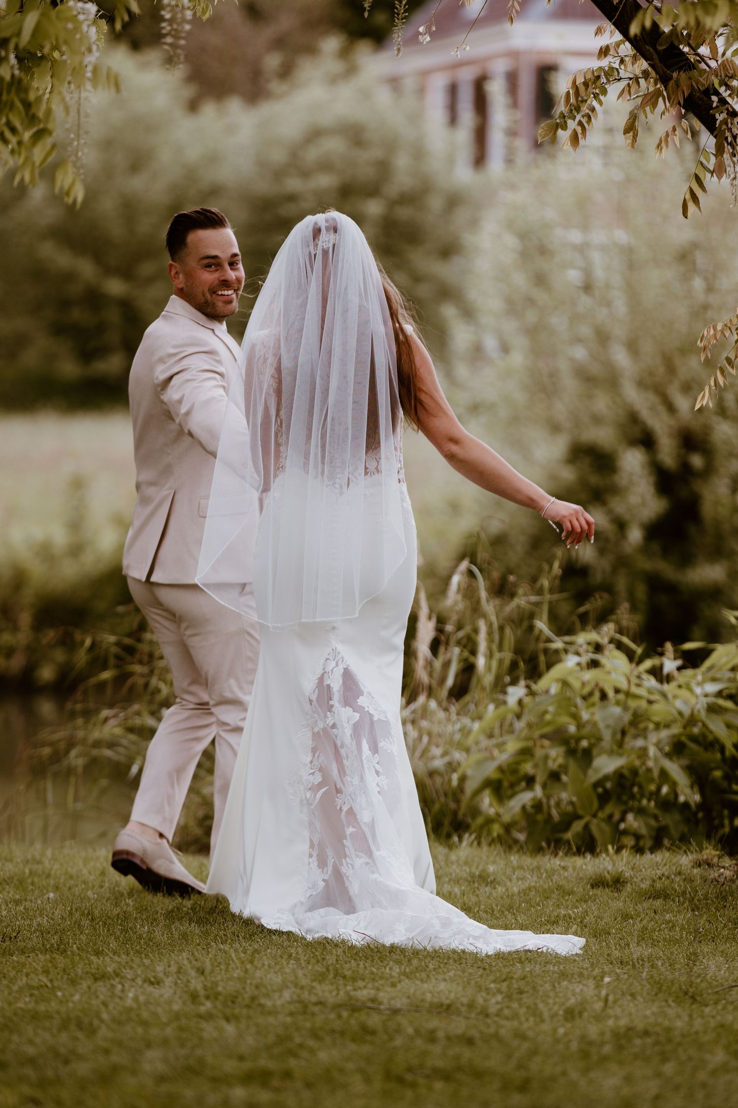 Groom smiles back at camera while walking with veiled bride through lush green garden.