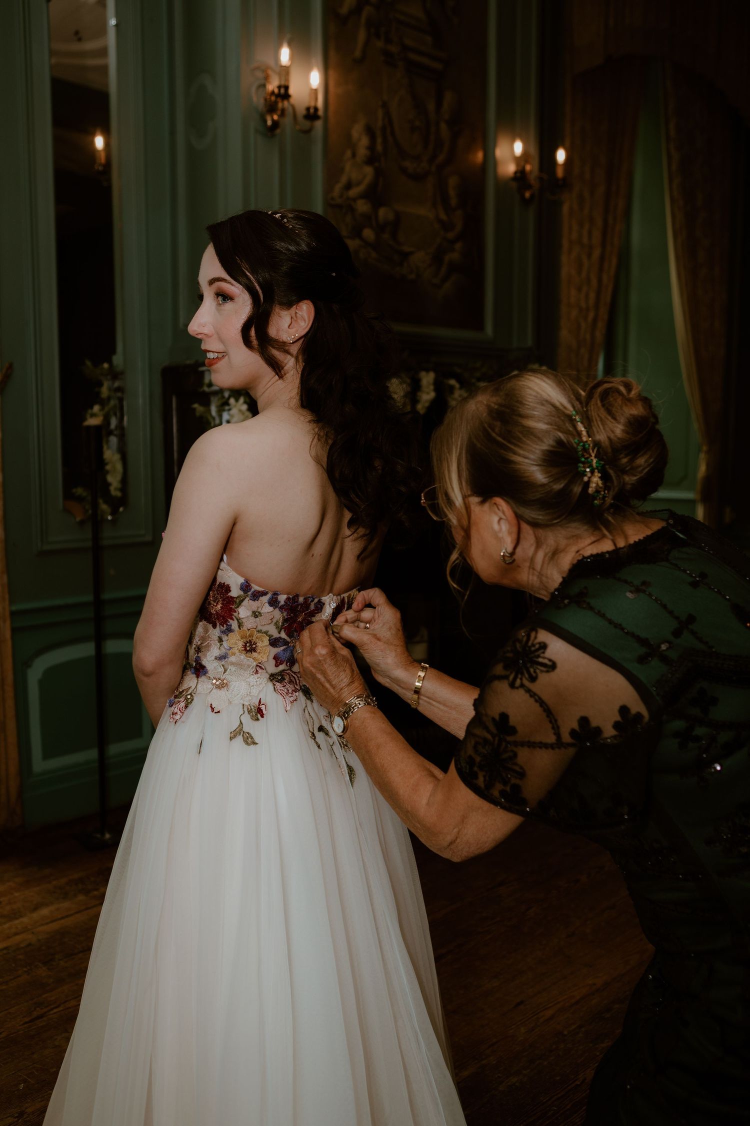 Bride in a floral embroidered backless gown being buttoned up by a woman in an ornate green room.