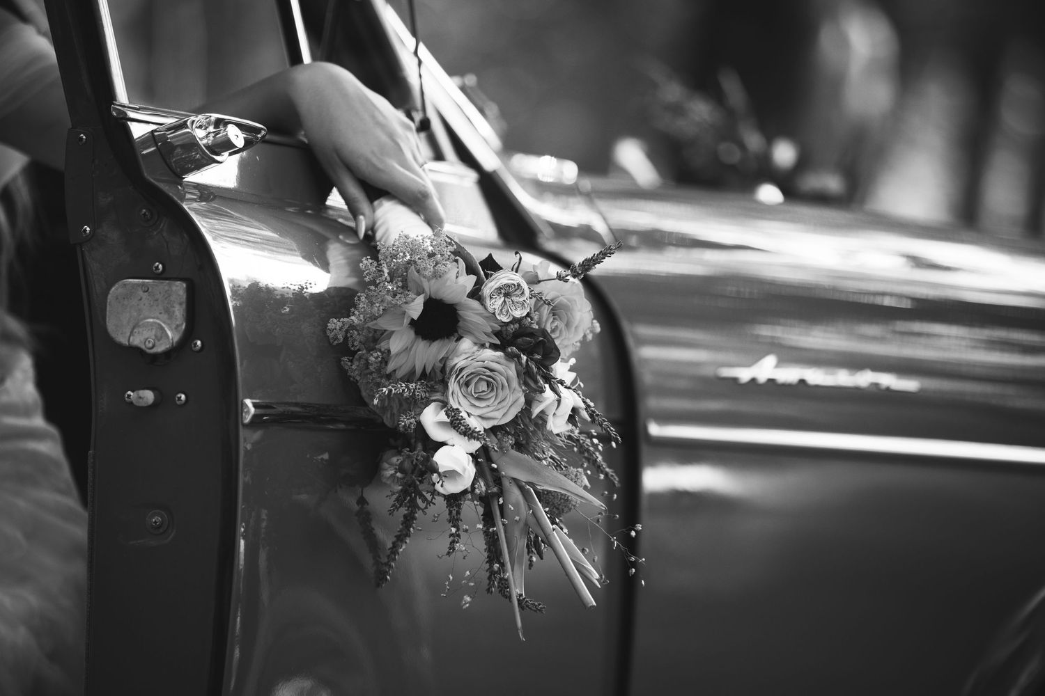 Black and white close-up of bridal bouquet resting on vintage classic car door.