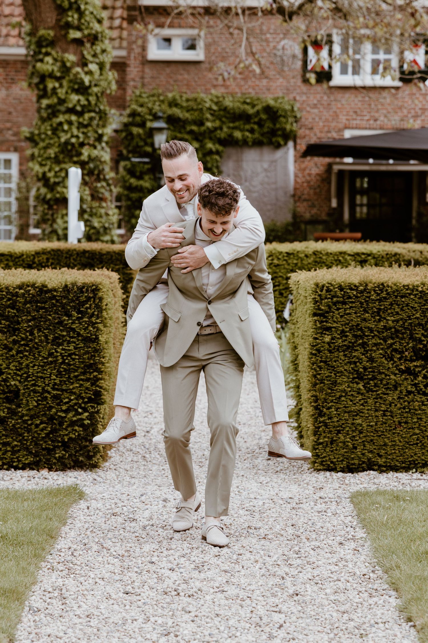 Two men in matching beige suits share a playful piggyback ride in a formal garden.