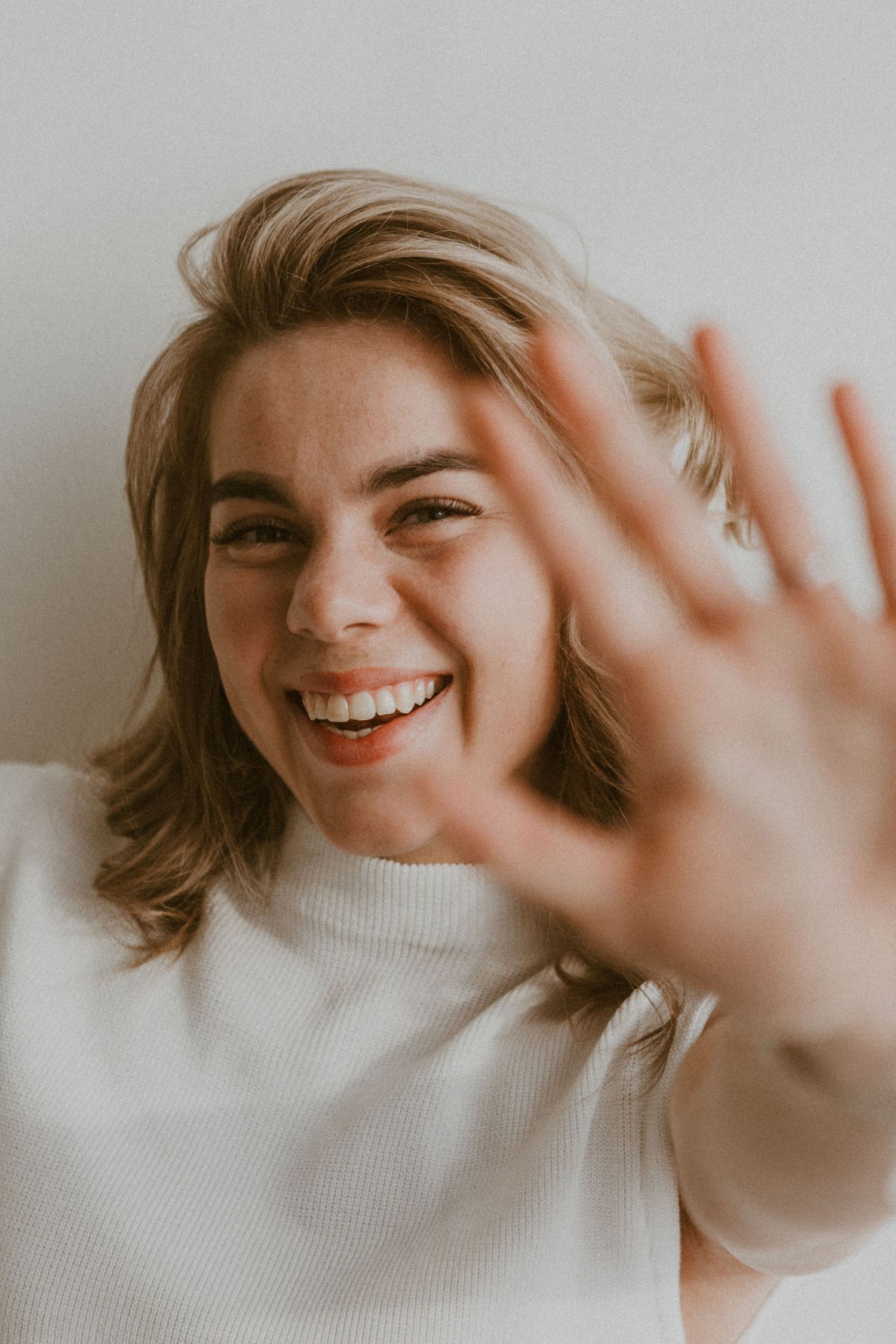 Cheerful blonde woman in a white turtleneck playfully reaching her hand toward the camera.
