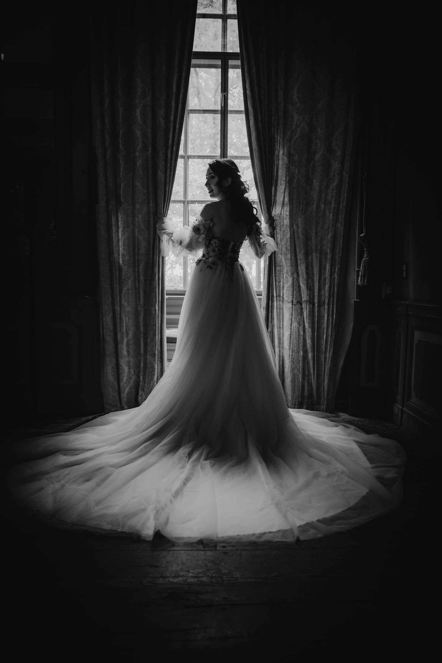 Black and white silhouette of bride in full ball gown with long train standing before grand window with curtains.