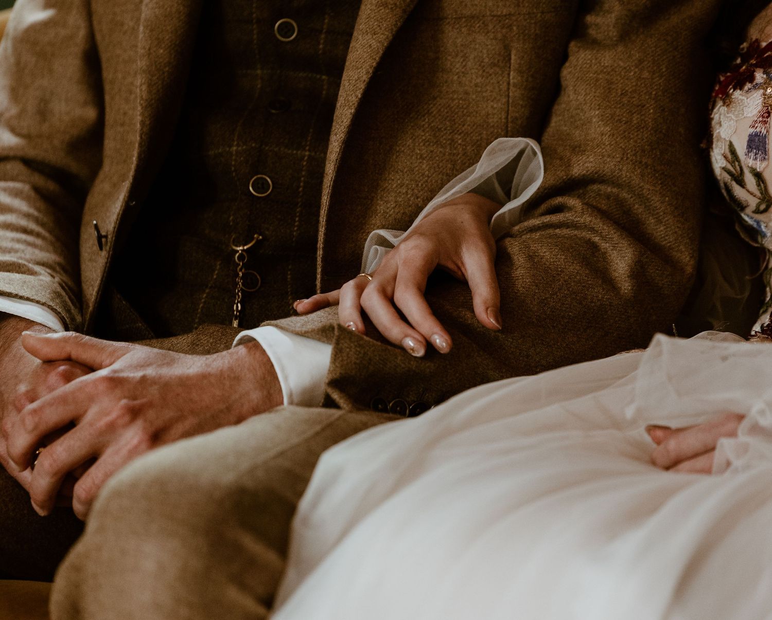 Close-up of bride and groom holding hands, groom in brown tweed suit with wedding ring visible.