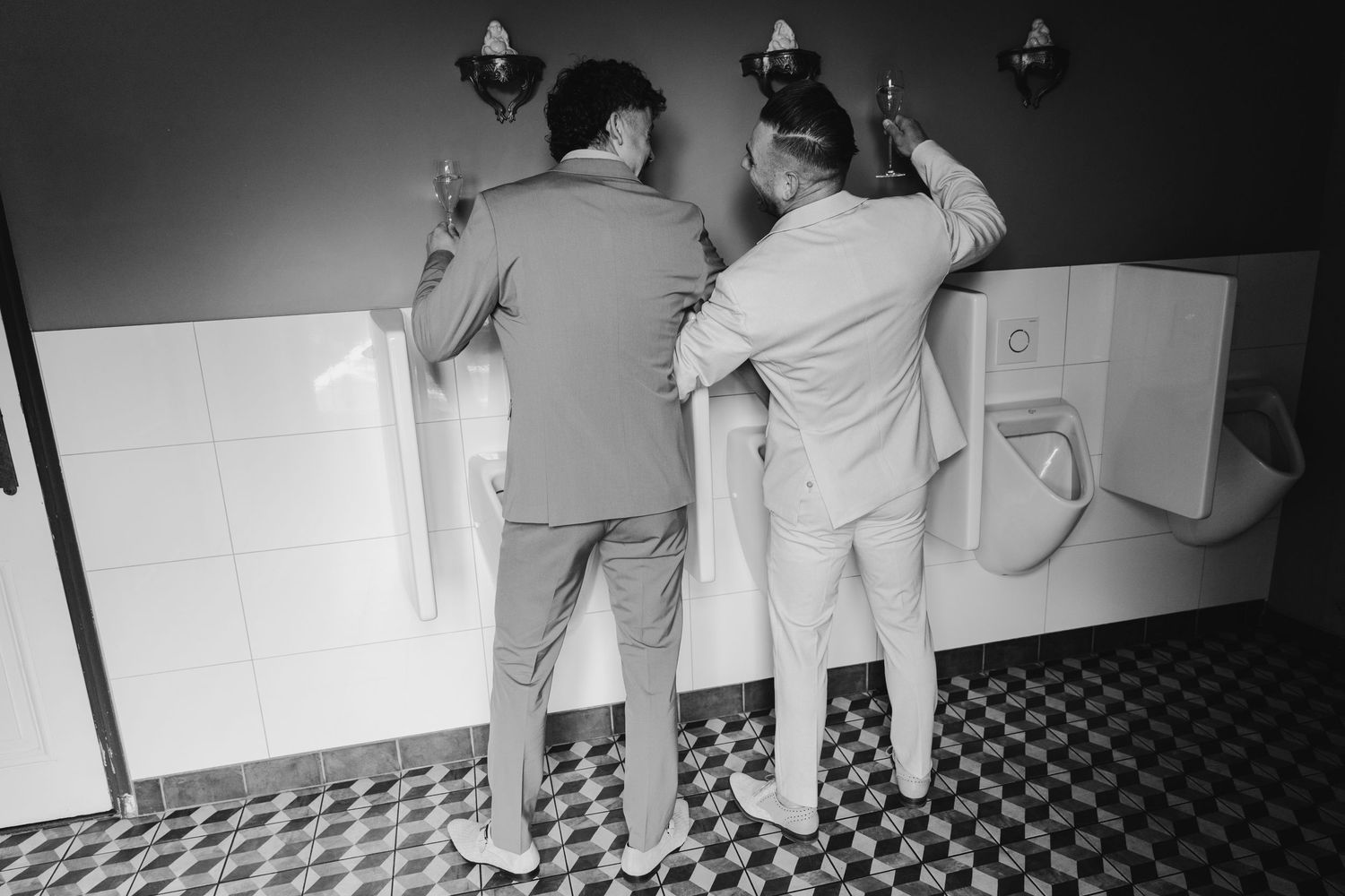Two groomsmen in suits stand together at urinals in a black and white wedding photo.