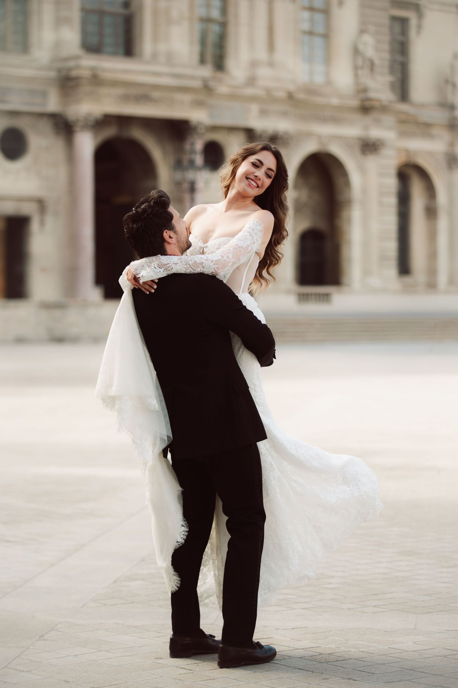 Groom lifts smiling bride in white lace wedding dress outdoors near a grand classical building in Paris.