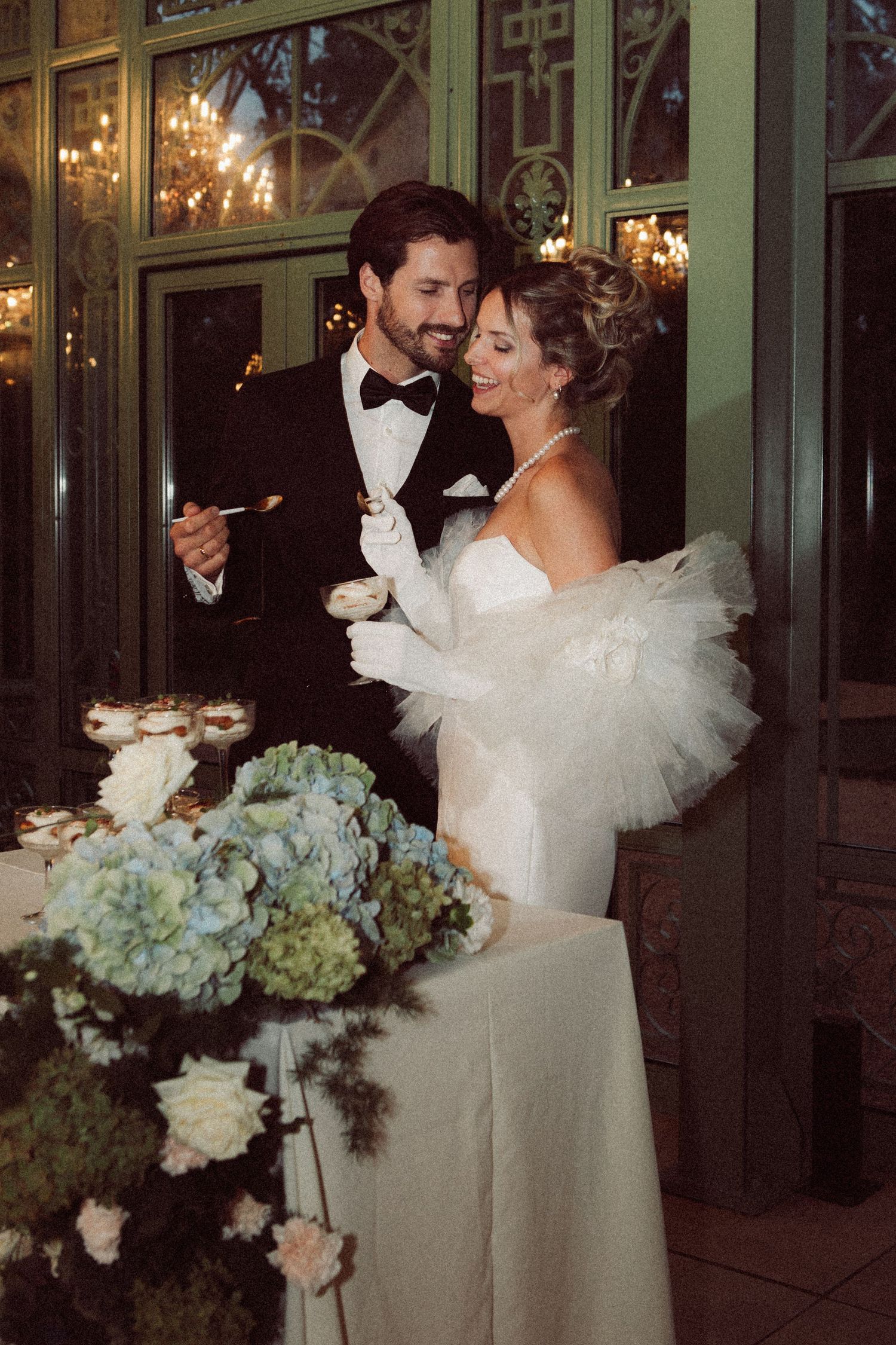 Elegant couple in wedding attire smiling while cutting cake, surrounded by blue hydrangea floral arrangements at reception.