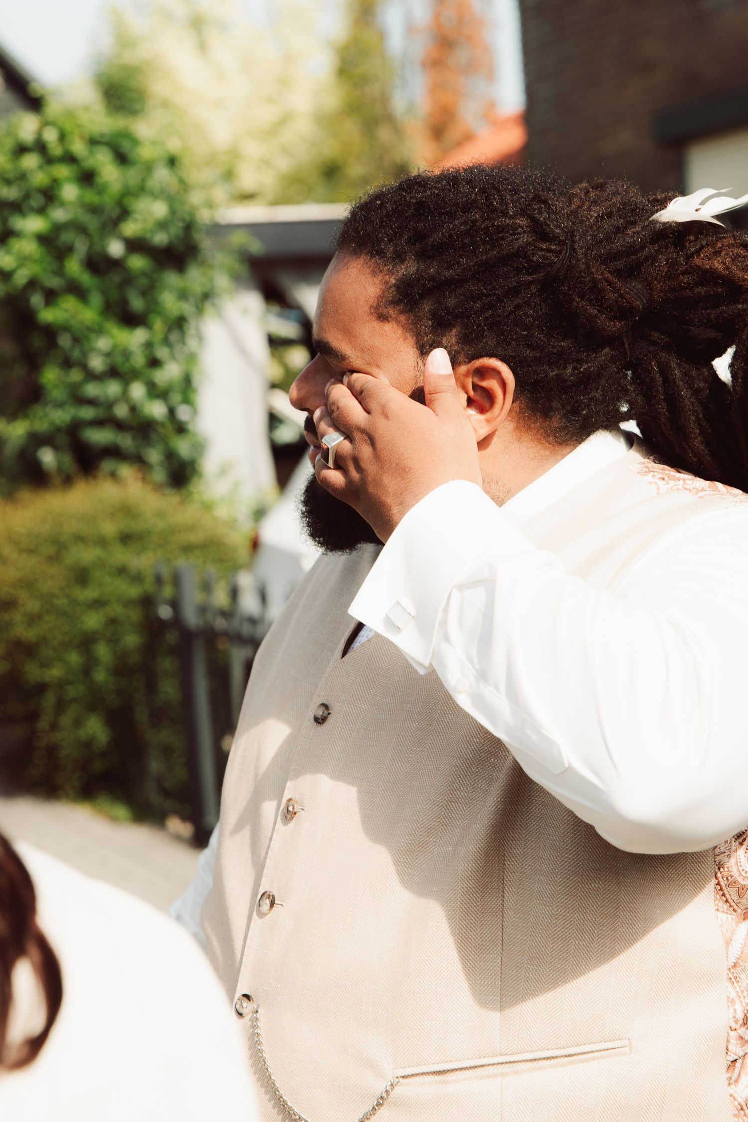 Emotional groom wiping tears on wedding day outdoors in sunlight.
