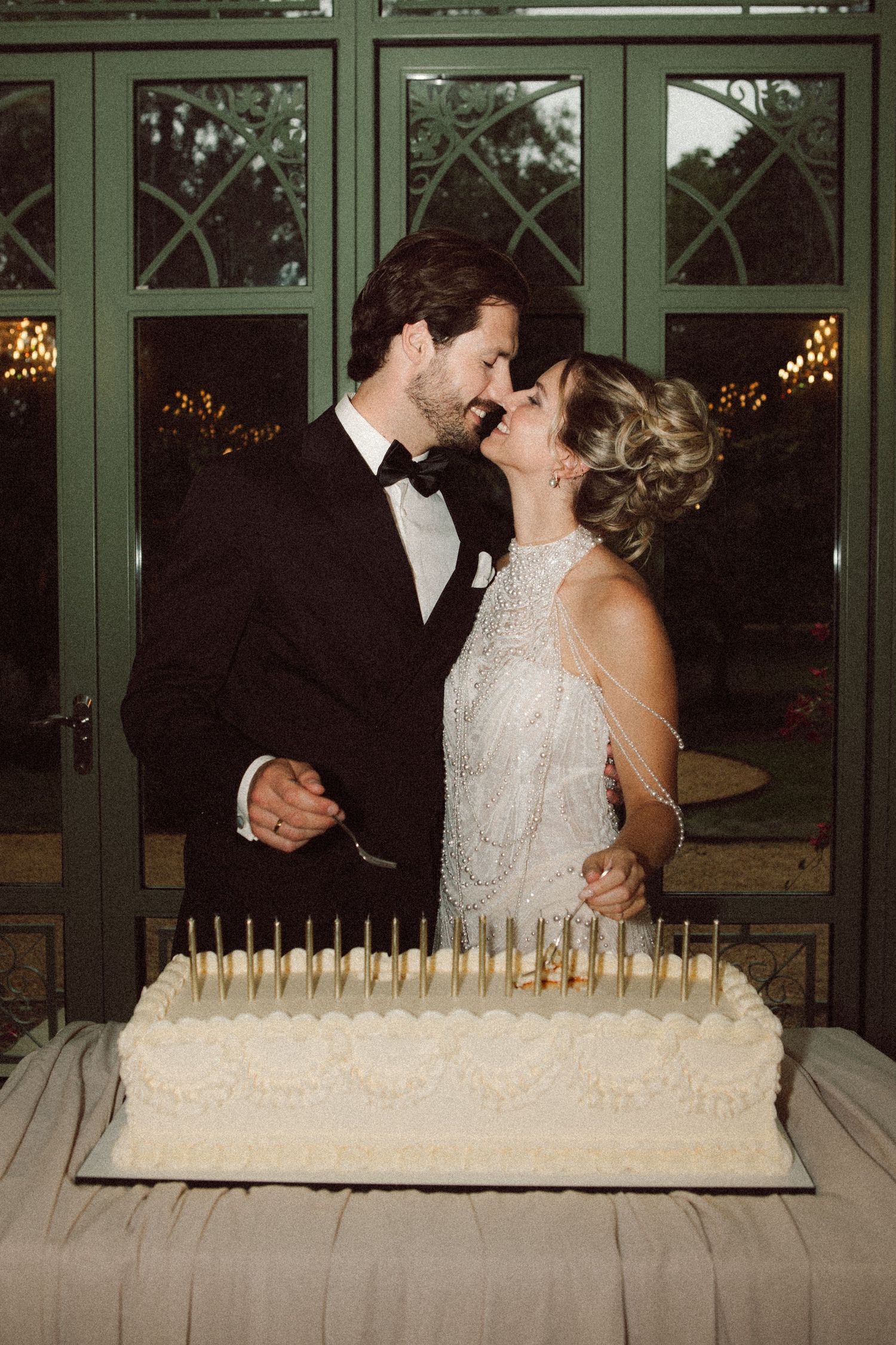 Bride and groom share a kiss while cutting their elegant white wedding cake adorned with gold candles.