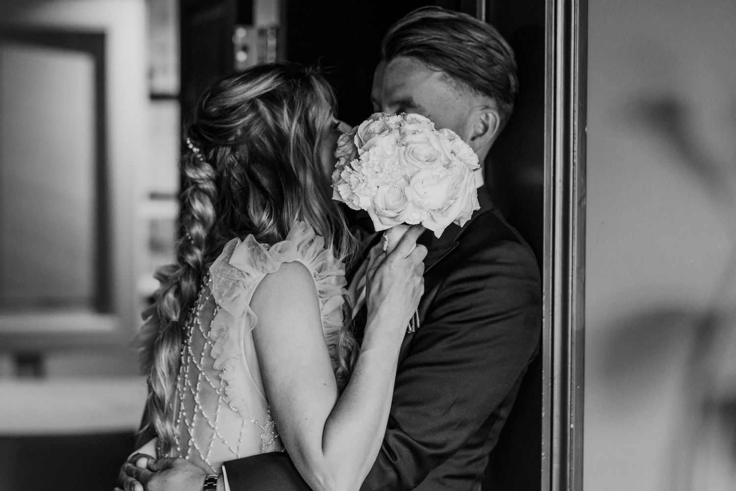 Black and white photo of bride and groom sharing a hidden kiss behind a white bridal bouquet.