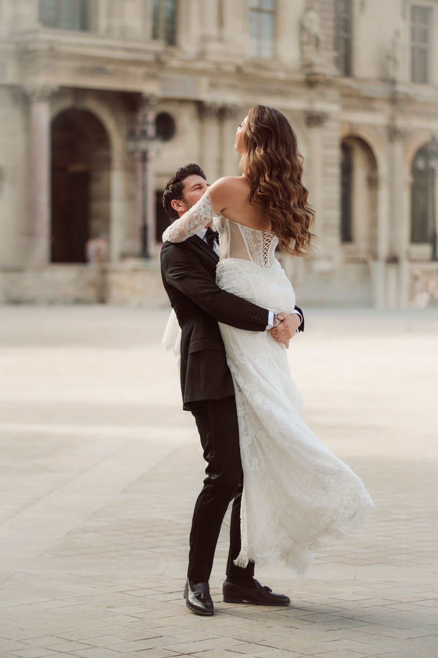 Groom lifts his bride in a flowing white lace gown in a sunlit Parisian courtyard.