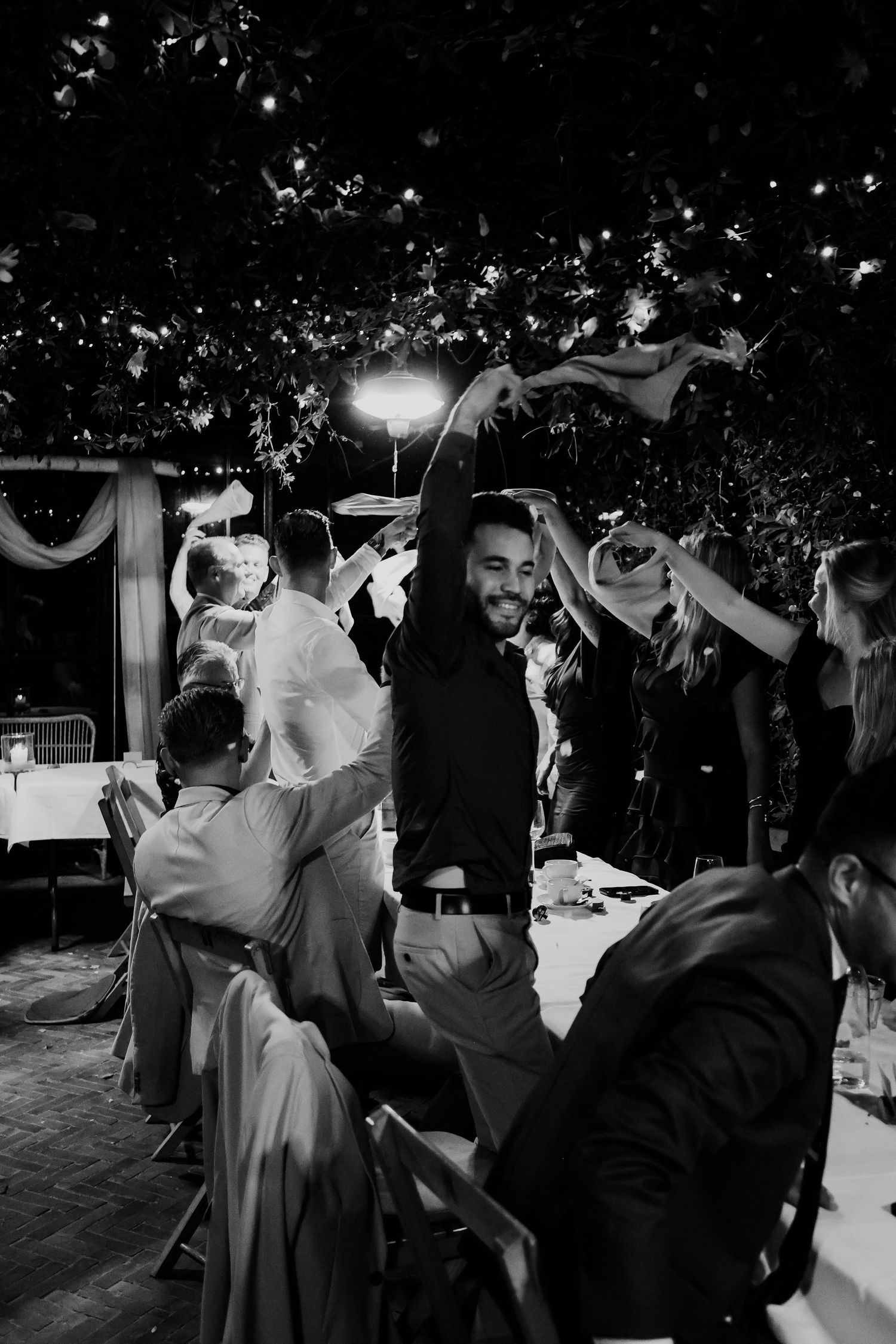 Joyful black and white wedding reception guests dancing and celebrating outdoors under string lights.