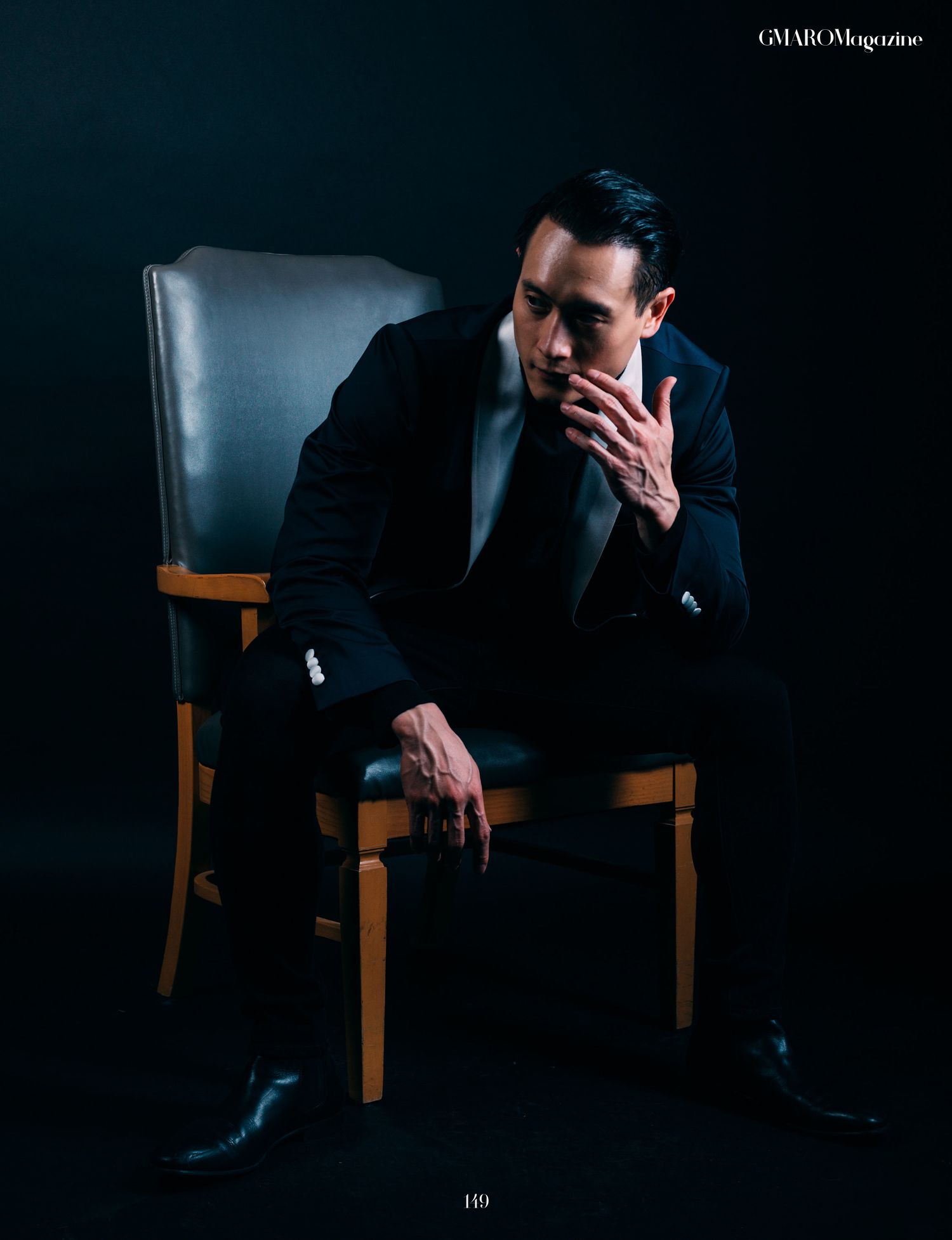 Contemplative studio portrait in dark tones showing figure seated in wooden armchair.