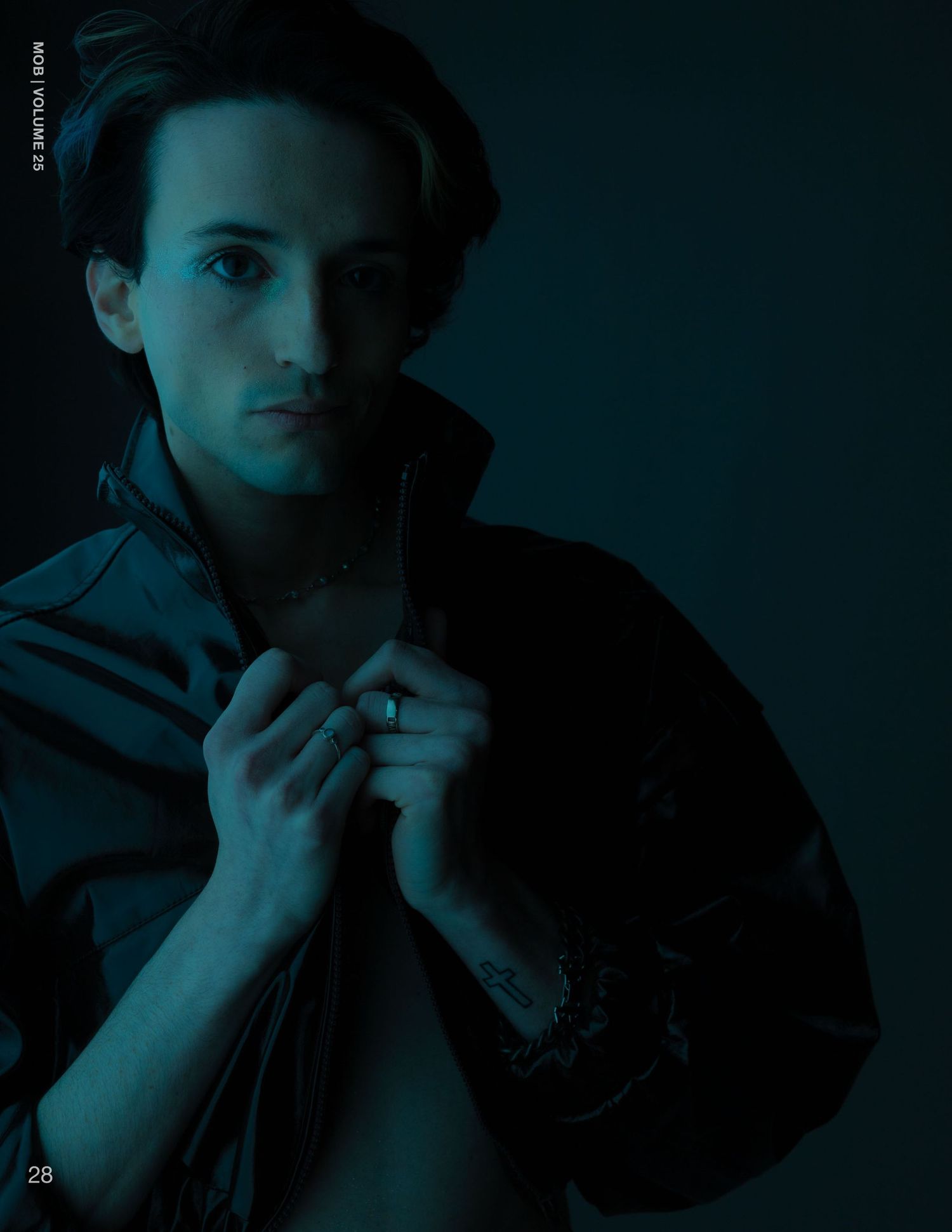 Young man in dark blue lighting poses with open jacket, wearing a necklace, in a moody editorial fashion shoot.