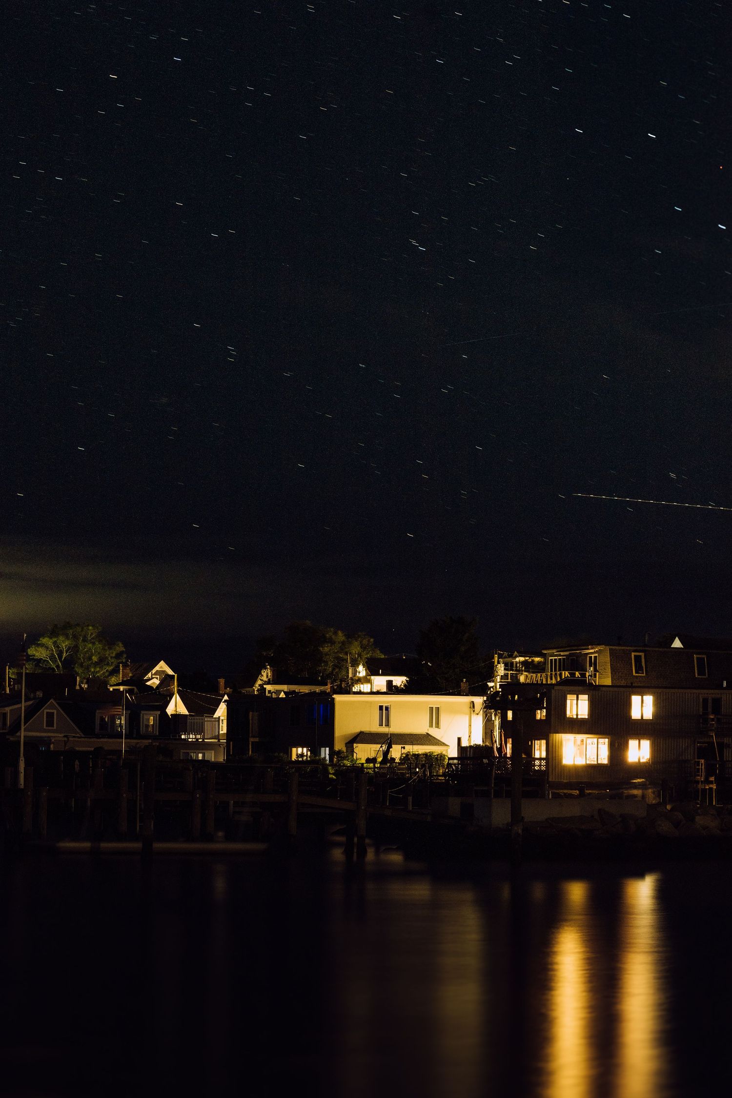 Nighttime waterfront village with glowing house lights reflecting on calm water under a star-filled dark sky.