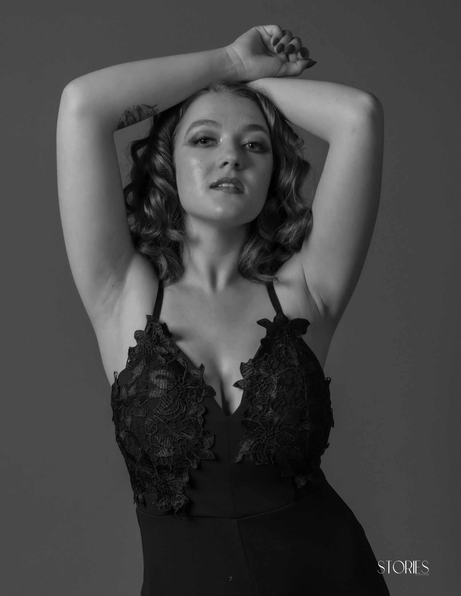 Black and white portrait of a young woman in lace bodysuit, arms raised gracefully above her head.
