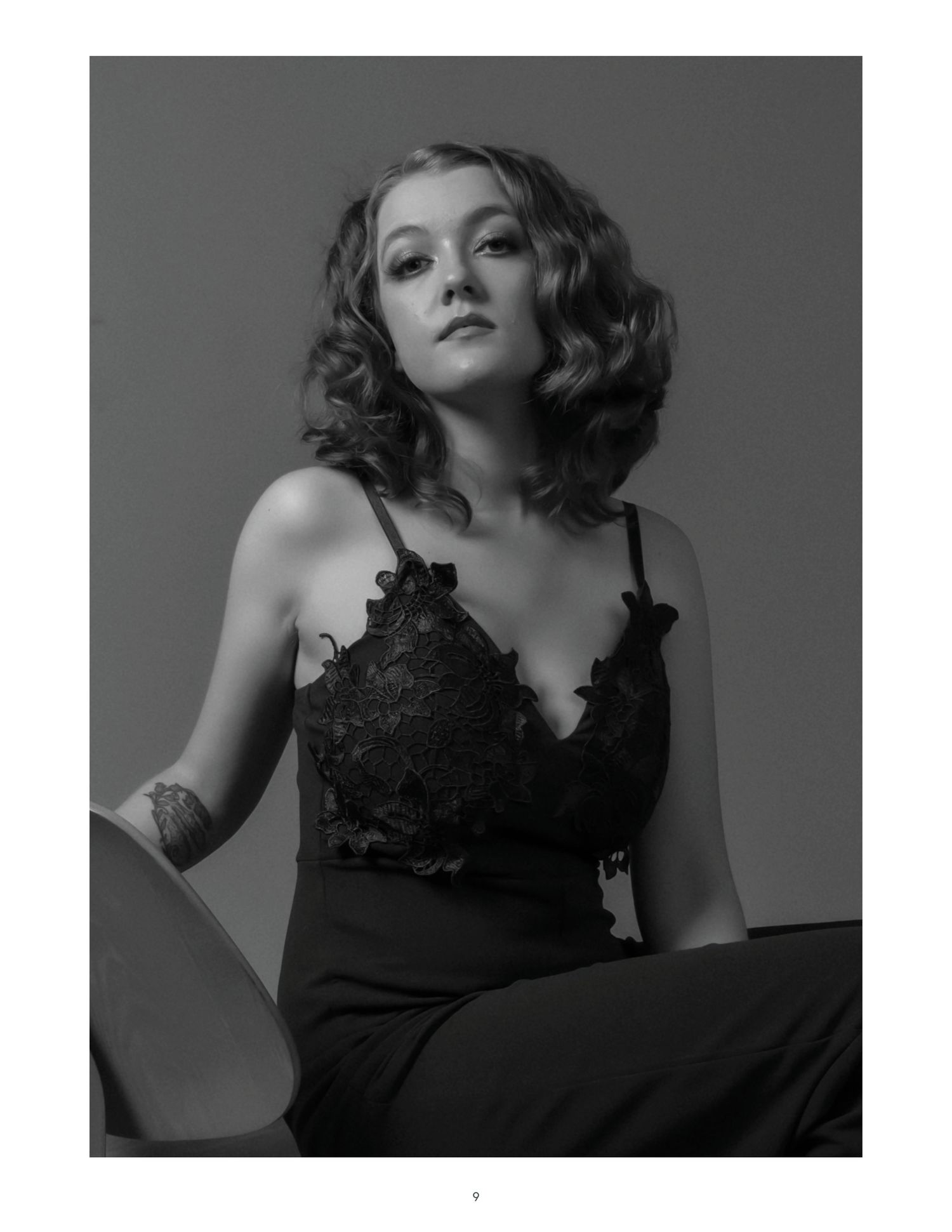 Black and white studio portrait of a young woman in lace top with curly hair and tattoo.