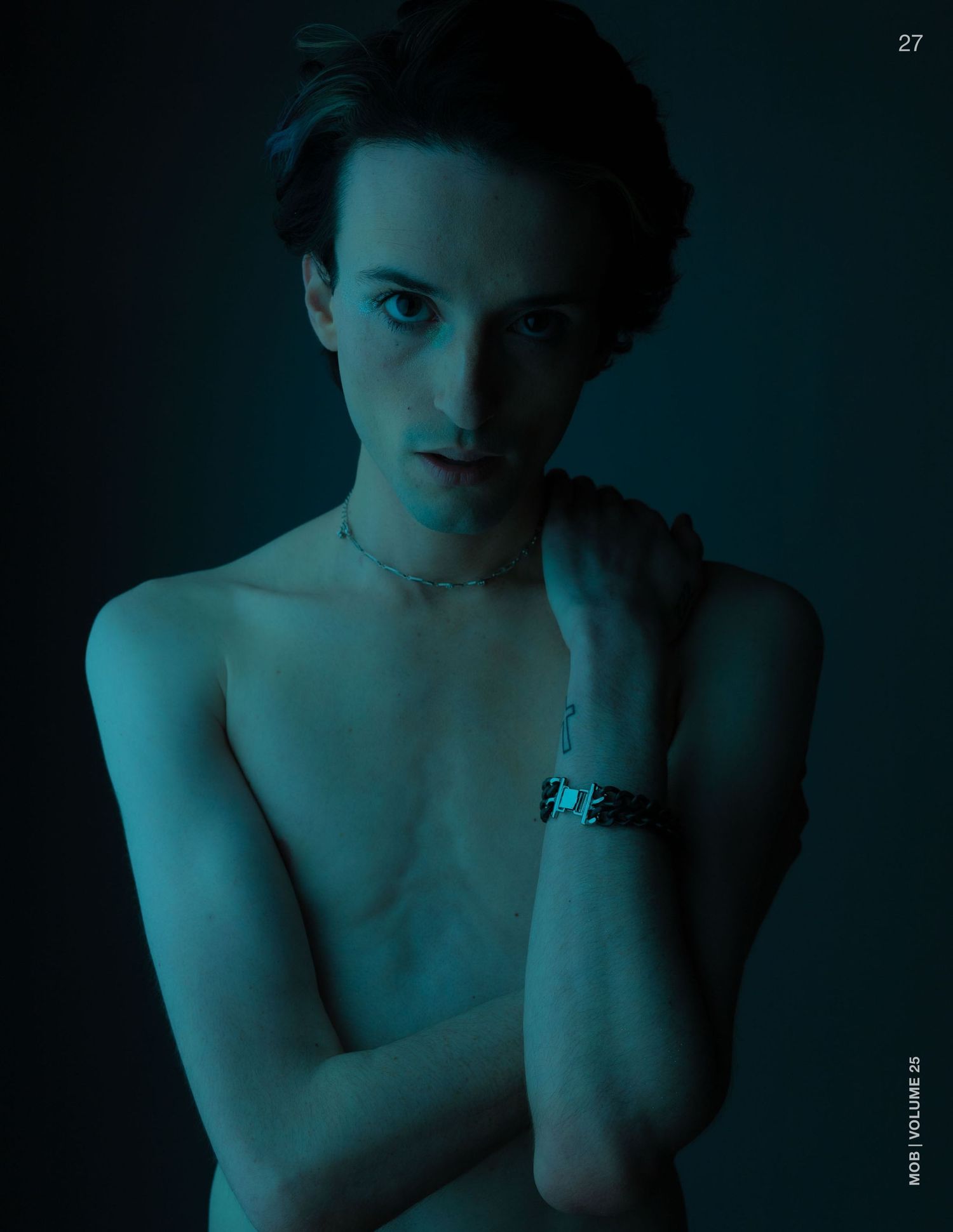 Shirtless male model with necklace and bracelet posing in dramatic blue-toned lighting.