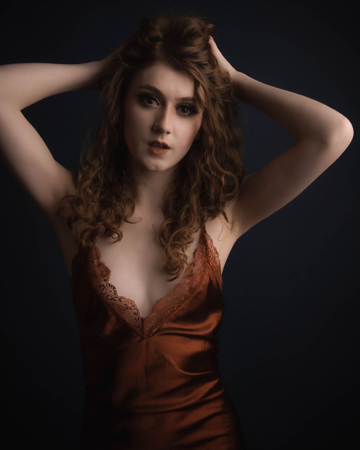 A woman with curly auburn hair poses in a rust-colored satin slip dress against a dark studio background.