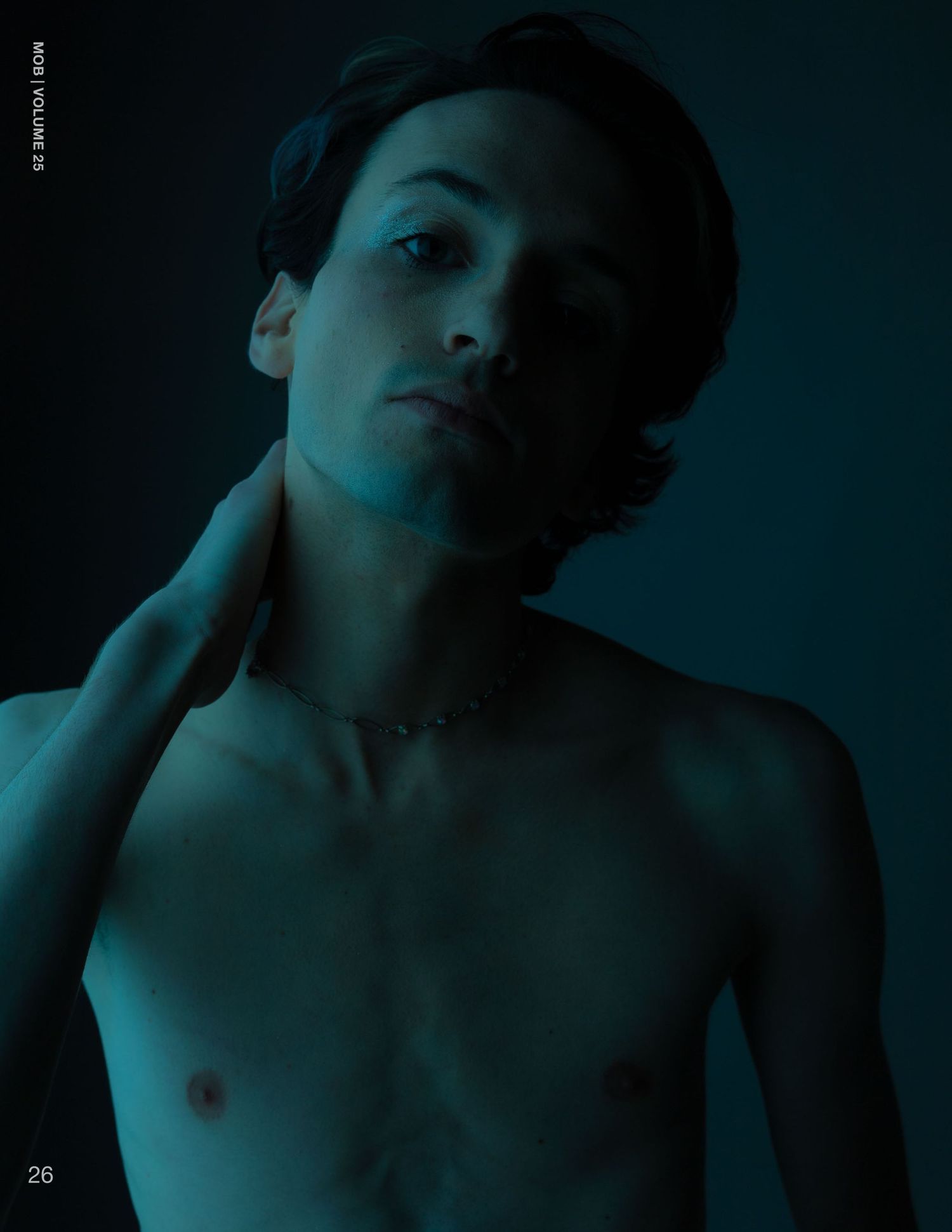 Shirtless male model wearing chain necklace poses dramatically in dark teal-lit editorial photo.