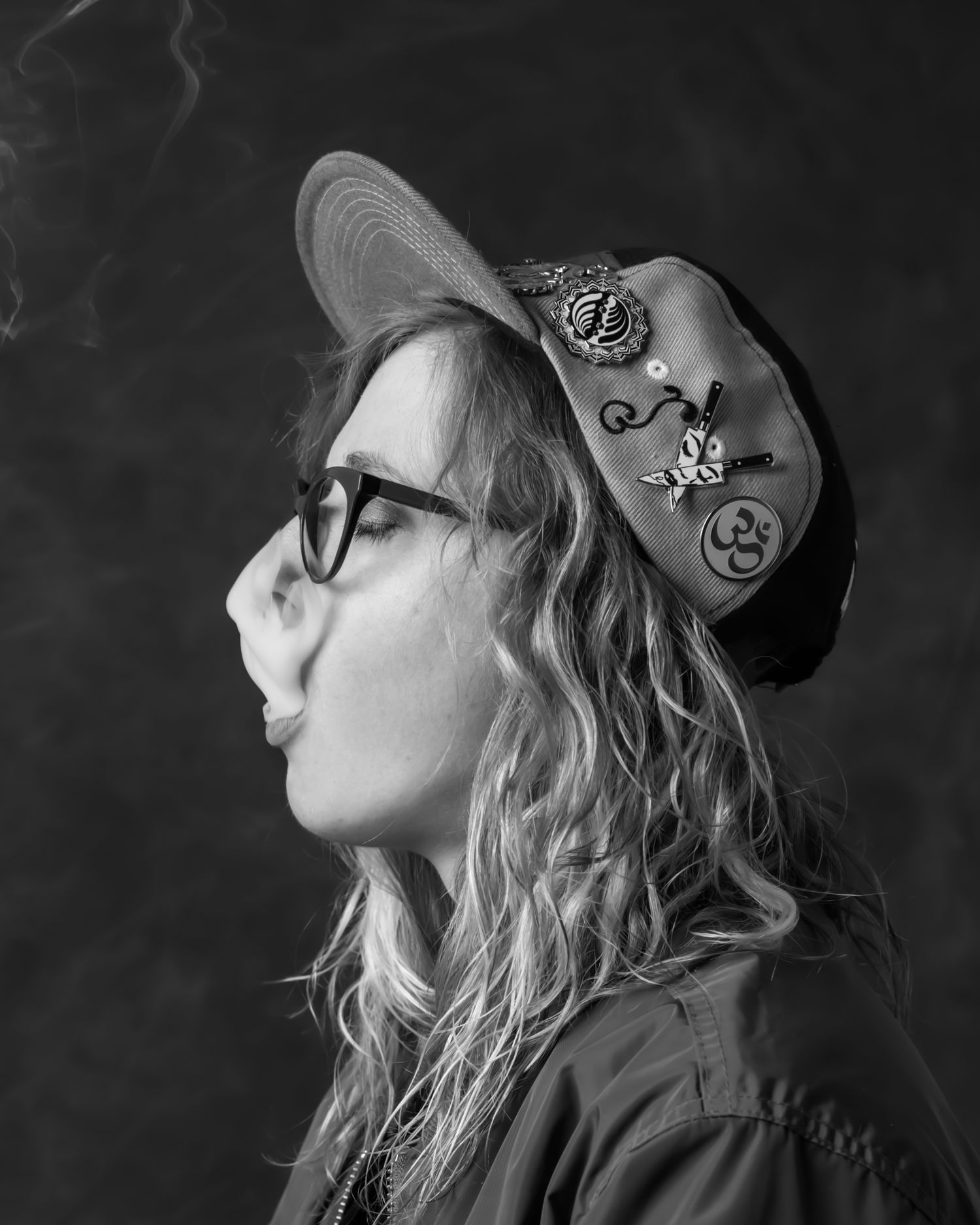 Side profile portrait of someone in decorated cap and glasses against dark background.