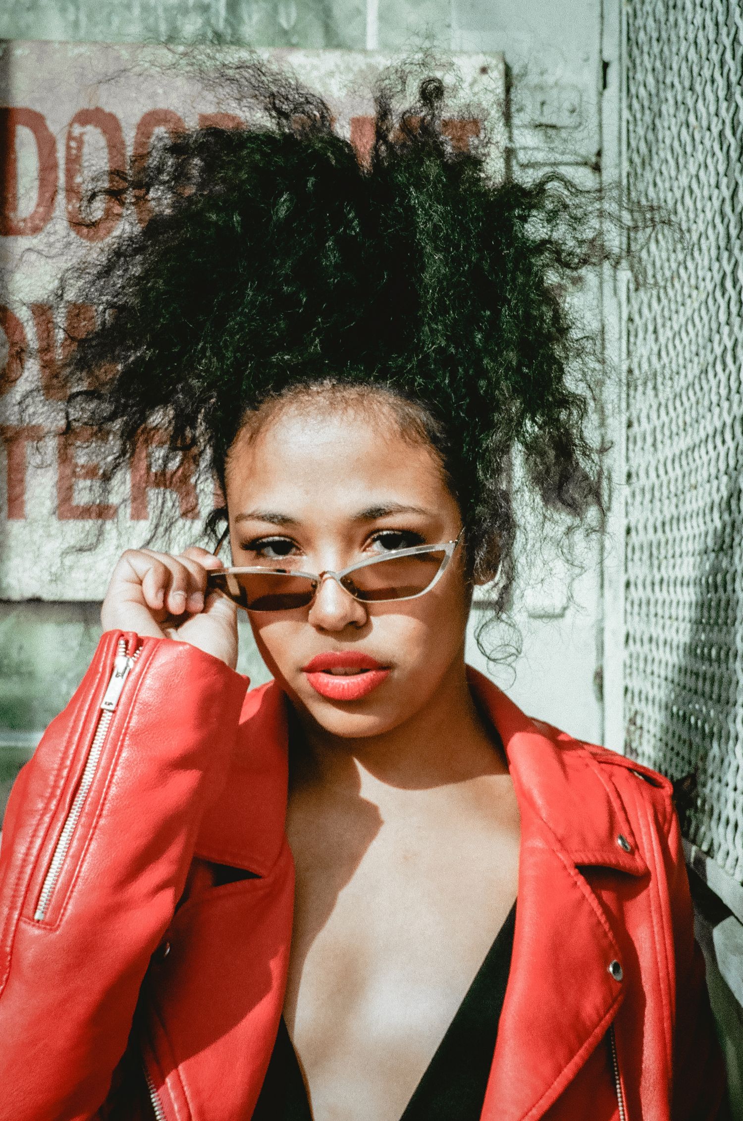 Fashion portrait featuring red leather jacket and retro sunglasses against urban backdrop.