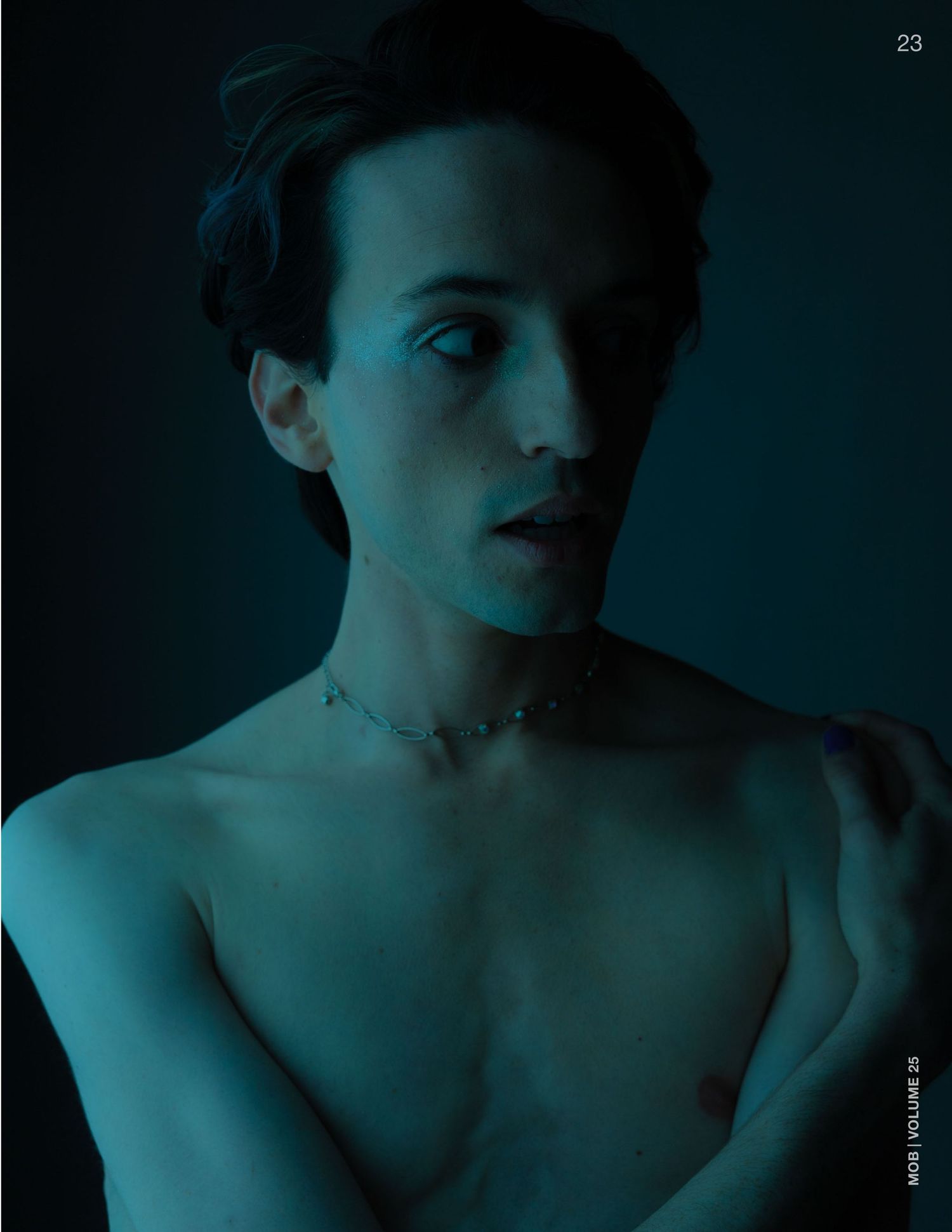 Shirtless model with a delicate chain choker necklace posed in dramatic blue-toned studio lighting.