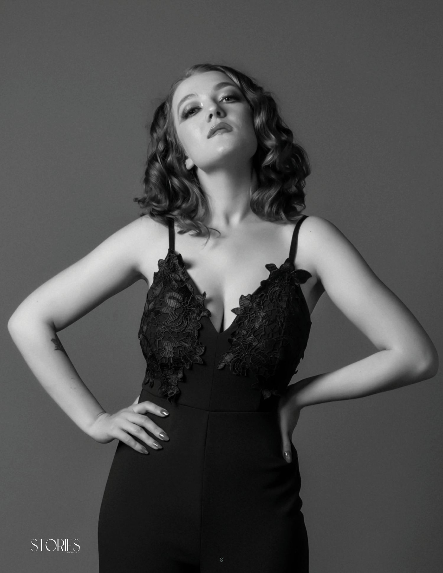 Black and white portrait of a woman in lace bodysuit with hands on hips, elegant vintage-style curled hair.