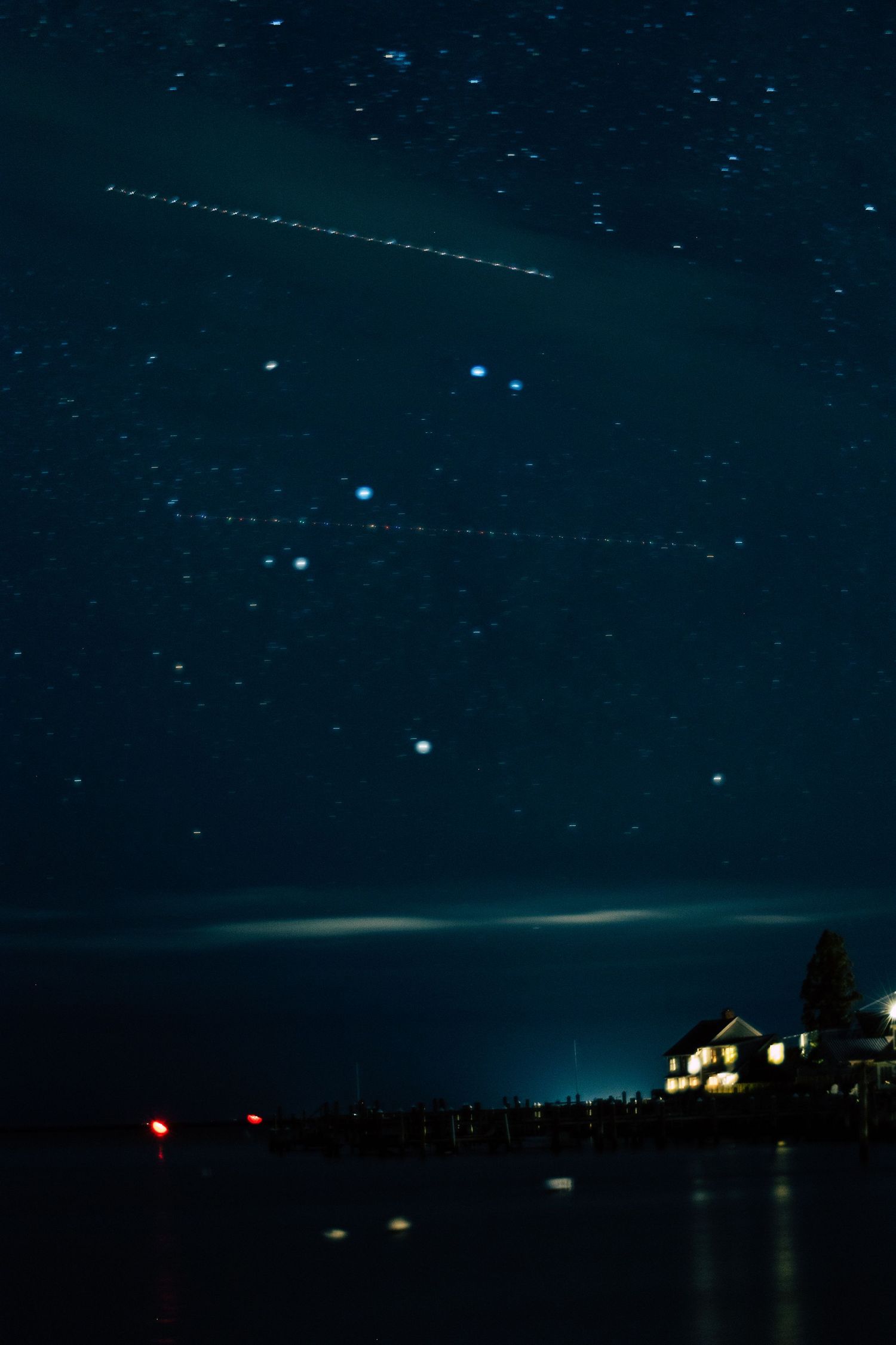 Starry night sky over a lake with illuminated pier and house, featuring a meteor streak and constellation.