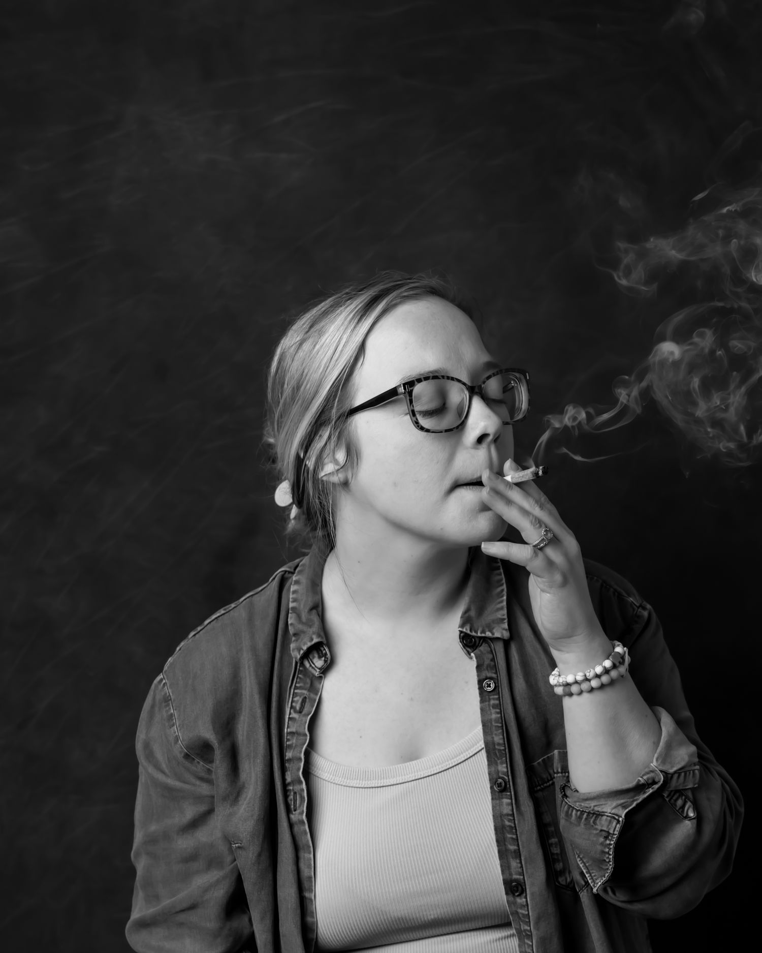 Black and white portrait of someone in glasses and denim jacket smoking.