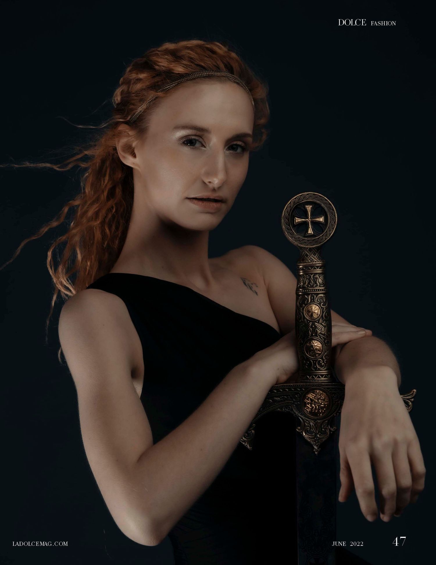 Red-haired woman in black dress holding ornate sword with cross guard, featured in Dolce Fashion magazine June 2022.