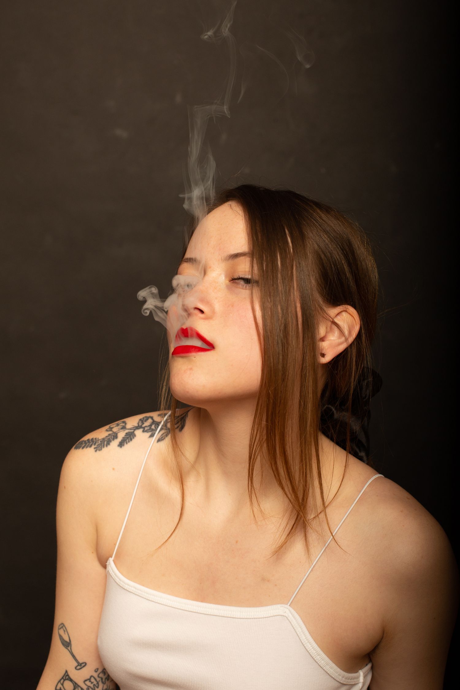 Artistic portrait of someone exhaling smoke while wearing a white top with visible tattoos against dark background.