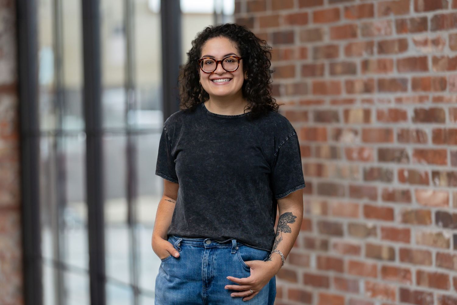 Smiling woman with curly hair and glasses in gray t-shirt and jeans, standing against a brick wall.