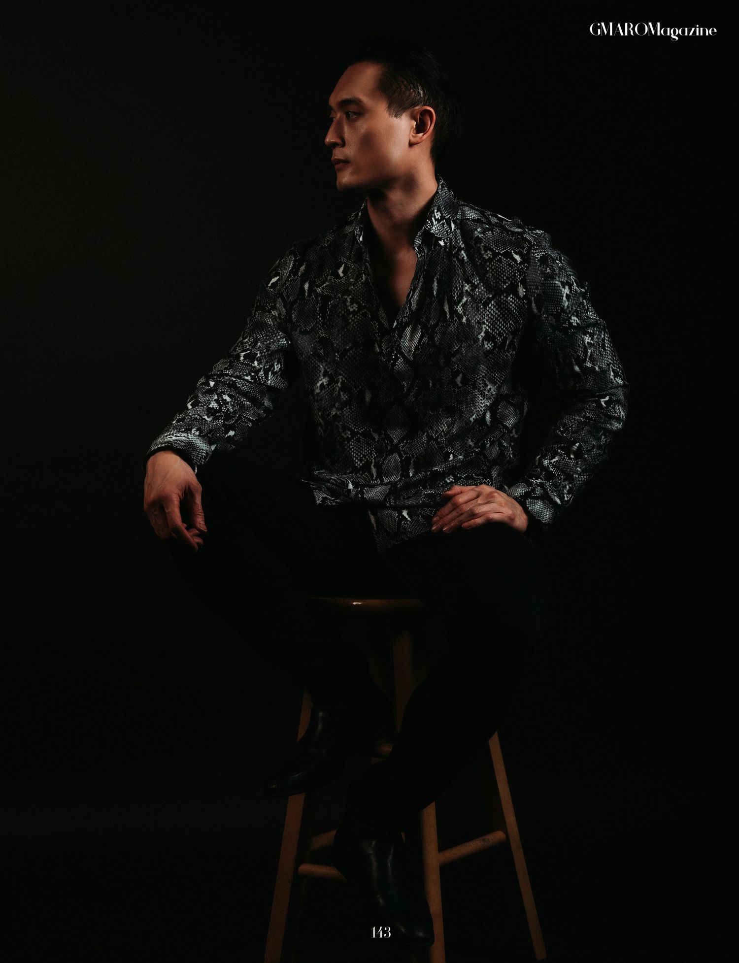 Editorial photos of model in patterned dark jacket seated on wooden stool.