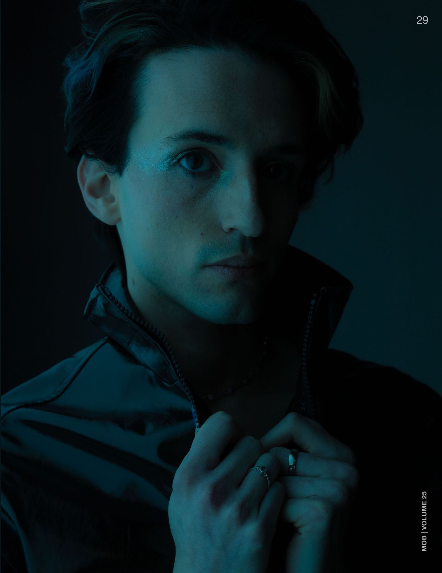 Young man in dark jacket poses dramatically under moody blue lighting in a stylish editorial portrait photo.