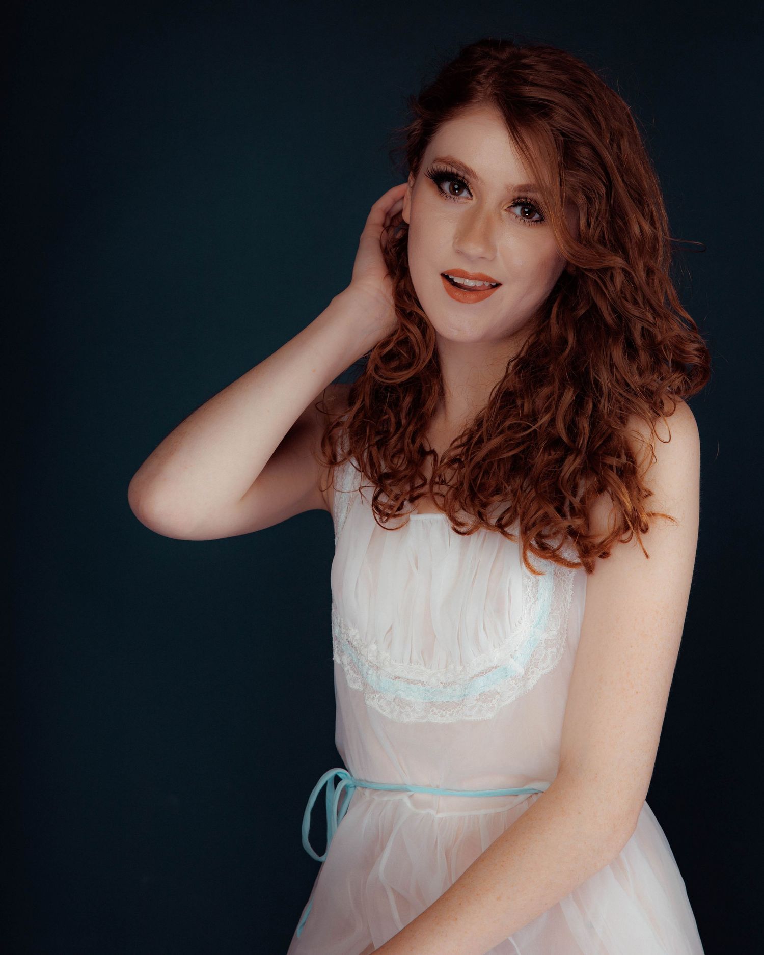Young woman with curly red hair poses gracefully in a white tulle dress with mint trim against dark background.