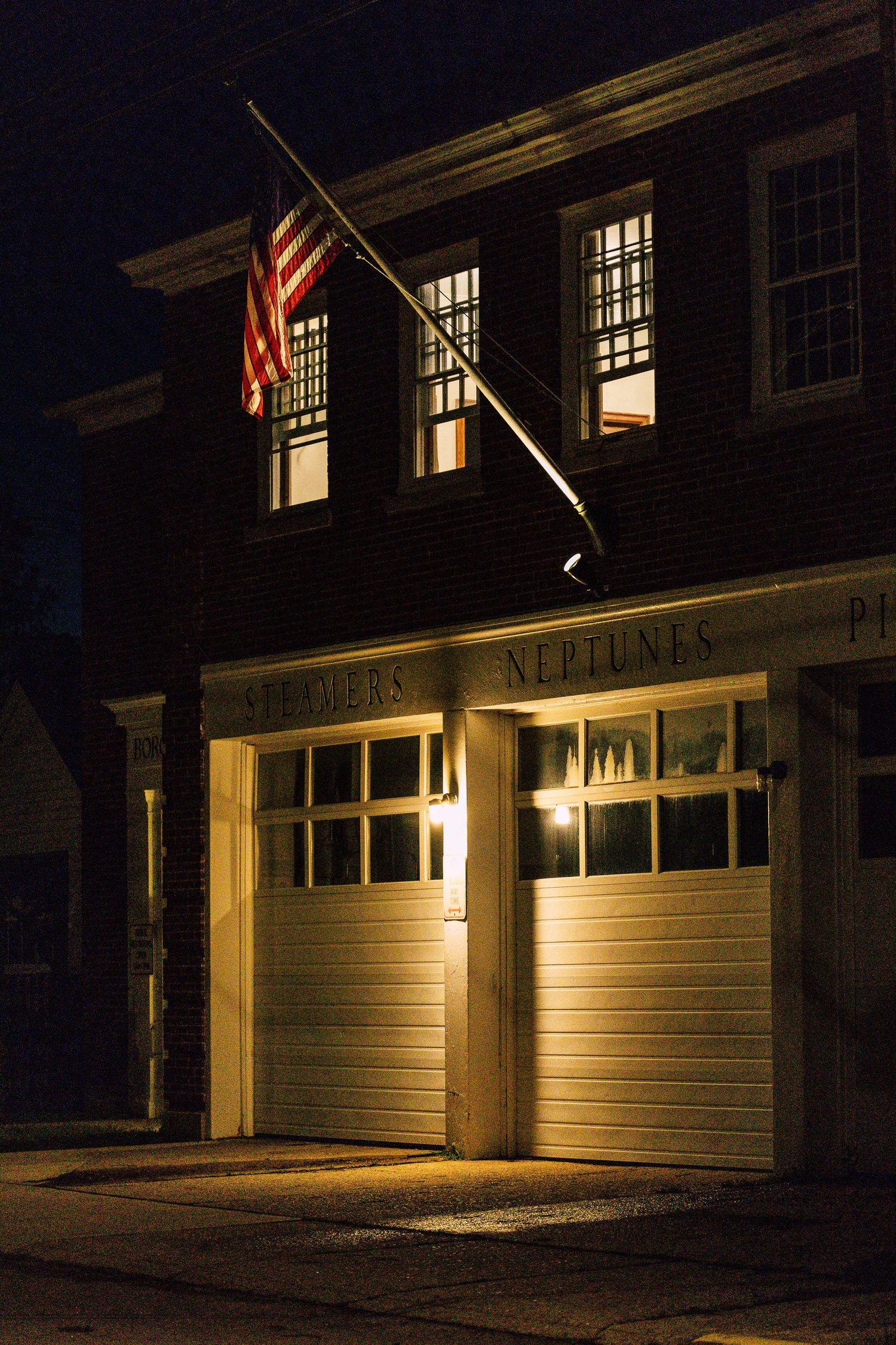Historic fire station at night with illuminated garage doors, American flag, and Neptune signage on a dark street.