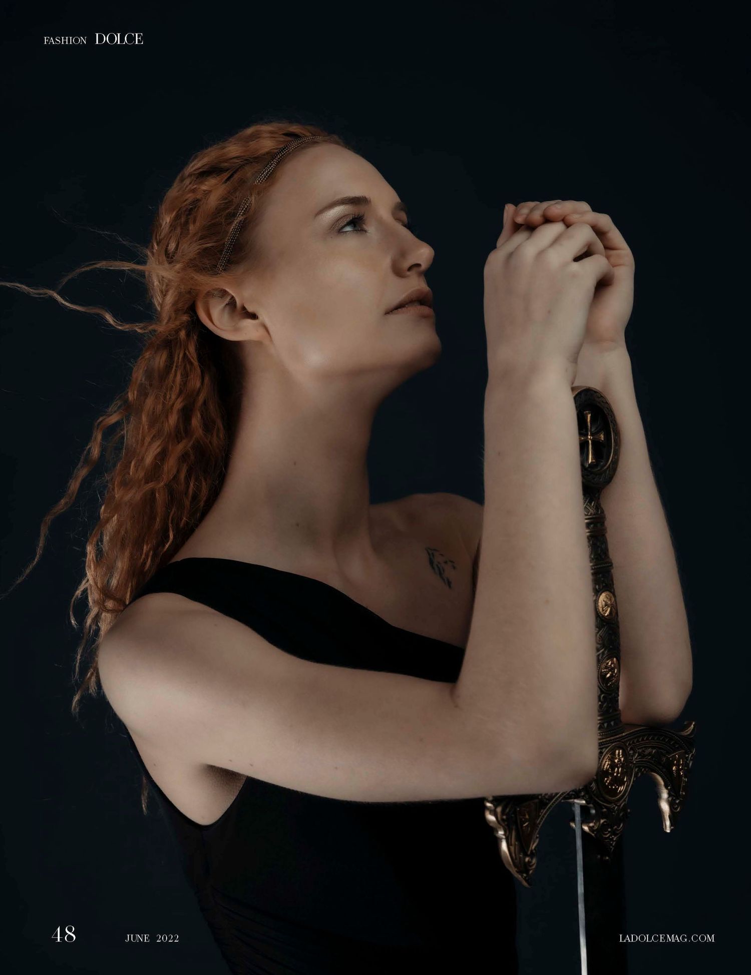 A red-haired woman in a black strapless top holds a ornate sword, gazing upward in a dramatic editorial pose.