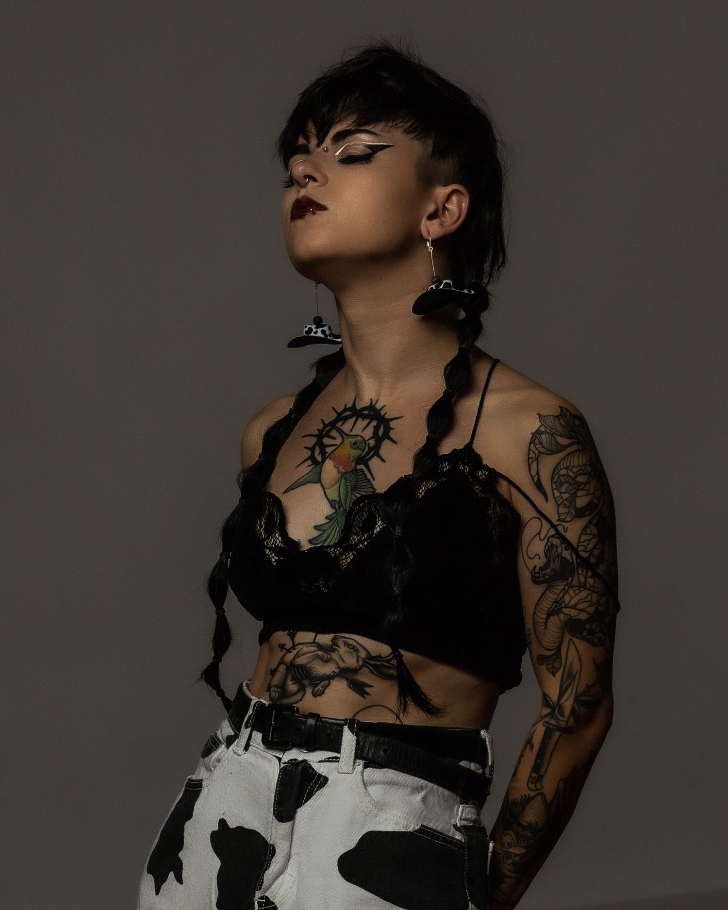 Tattooed woman in black lace crop top and cow-print pants poses with eyes closed, showcasing arm and chest tattoos.