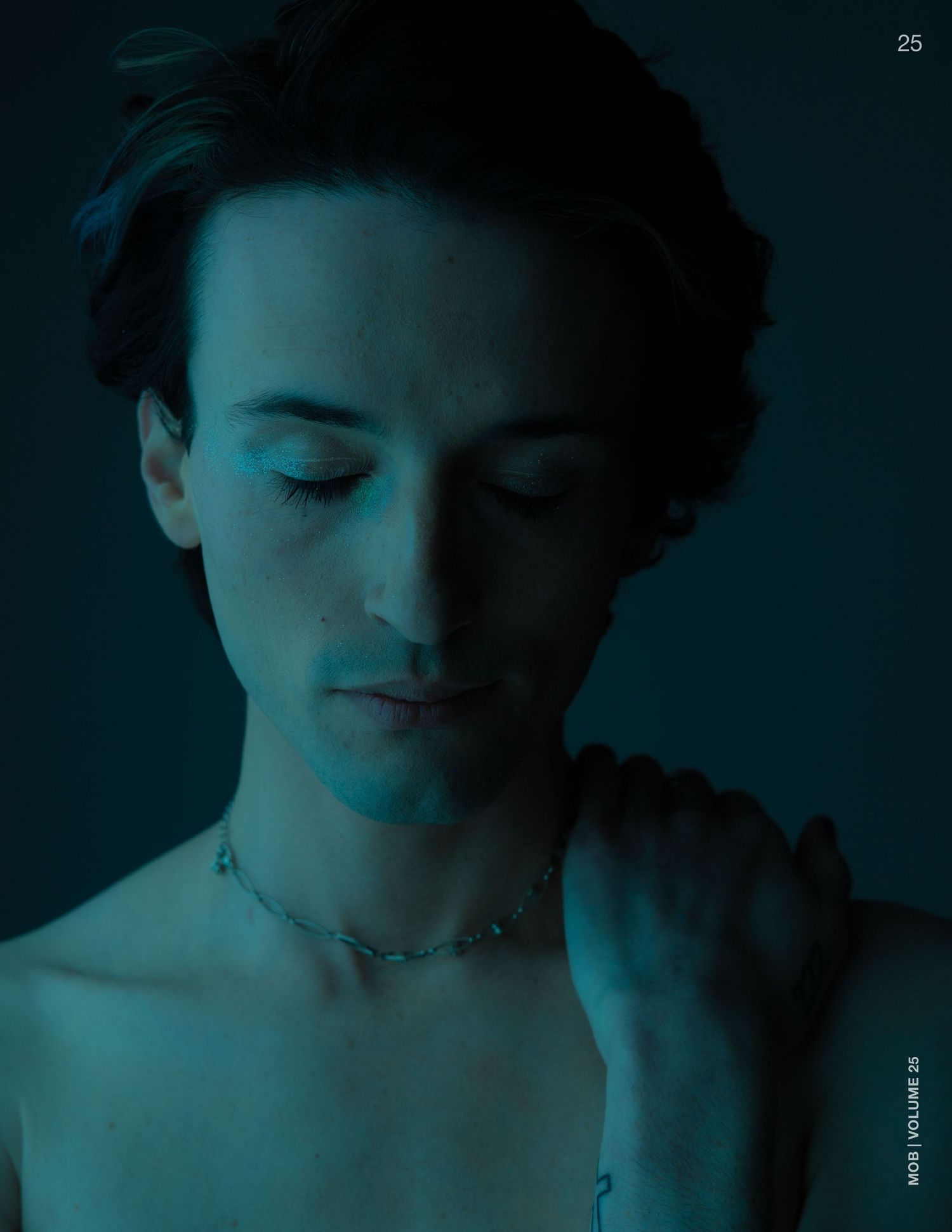 Person with closed eyes and a delicate chain necklace, bathed in moody blue-teal light.