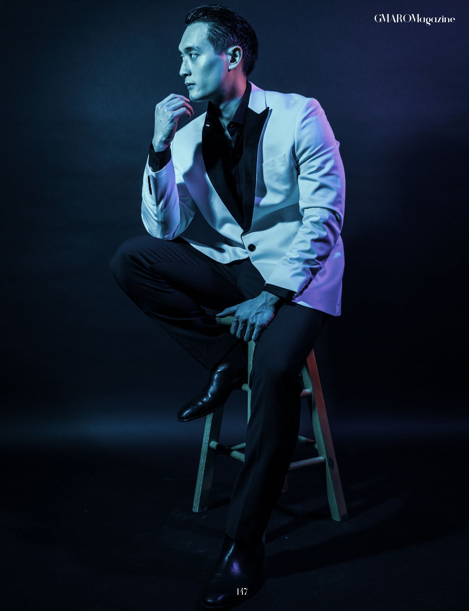 Artistic blue-lit studio photo of model in light blazer posed on wooden stool.