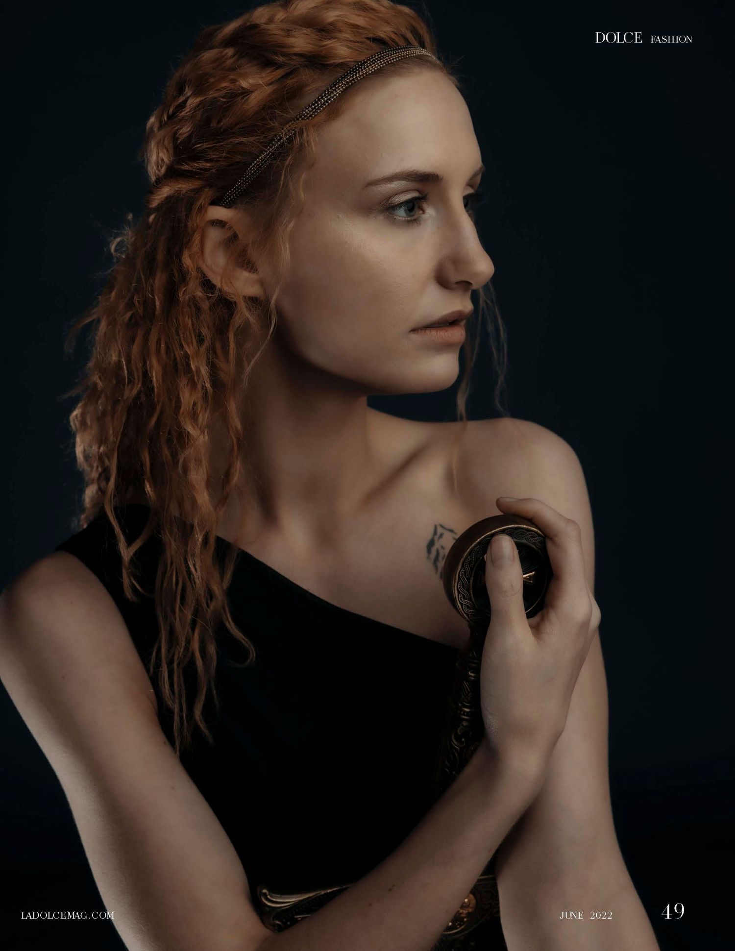 Woman with red braided hair in black off-shoulder top holds gold bracelet, featured in Dolce Fashion magazine.