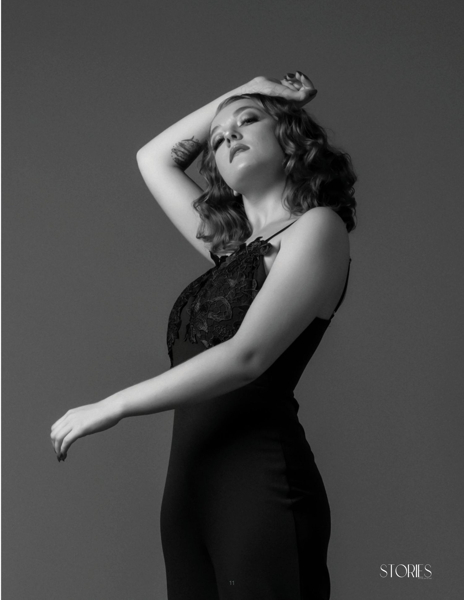 Black and white fashion portrait of a woman in elegant black dress posing dramatically against a studio background.